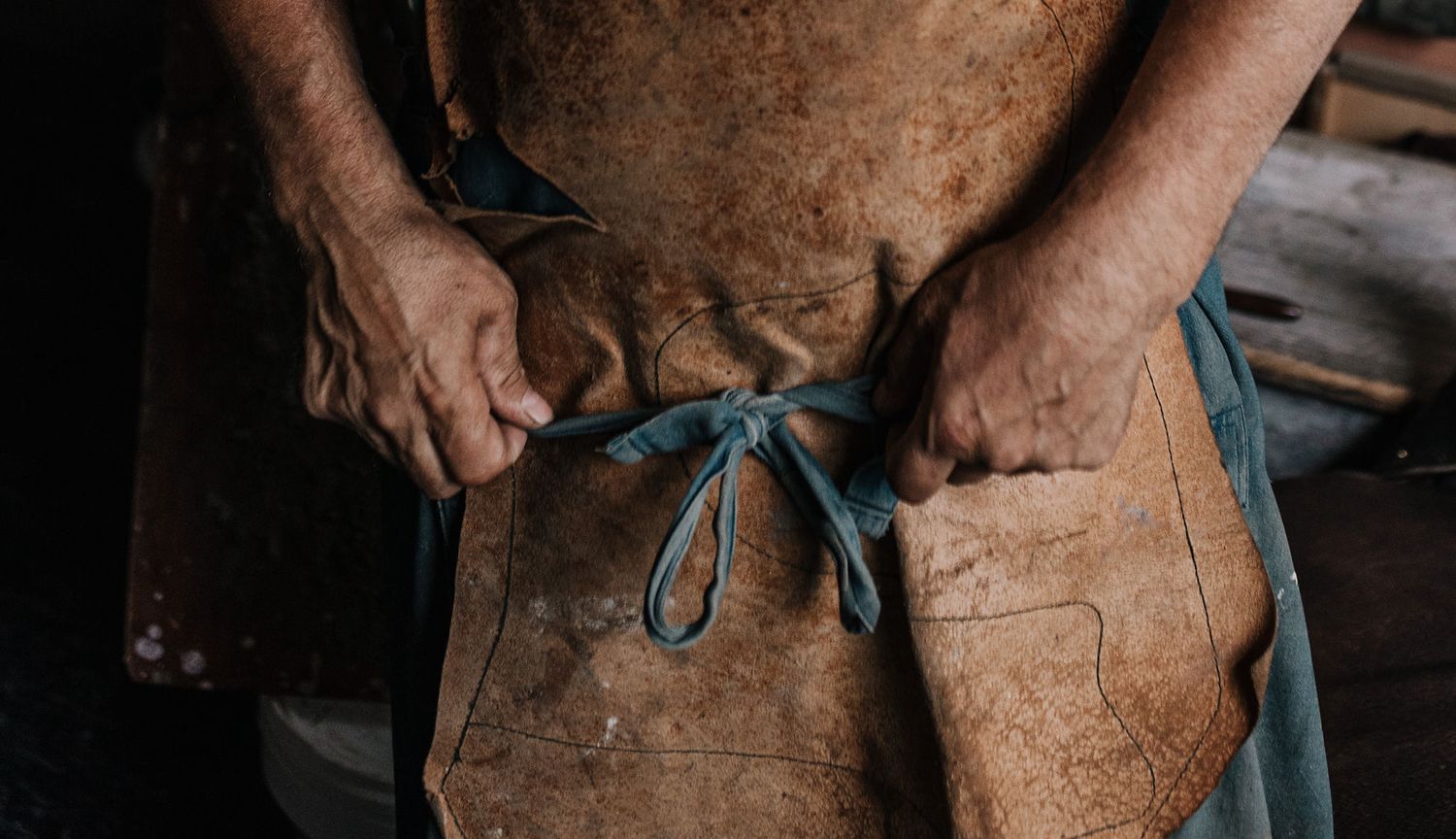 Weathered hands in a workshop wearing a brown leather apron covered in creative marks and stains.