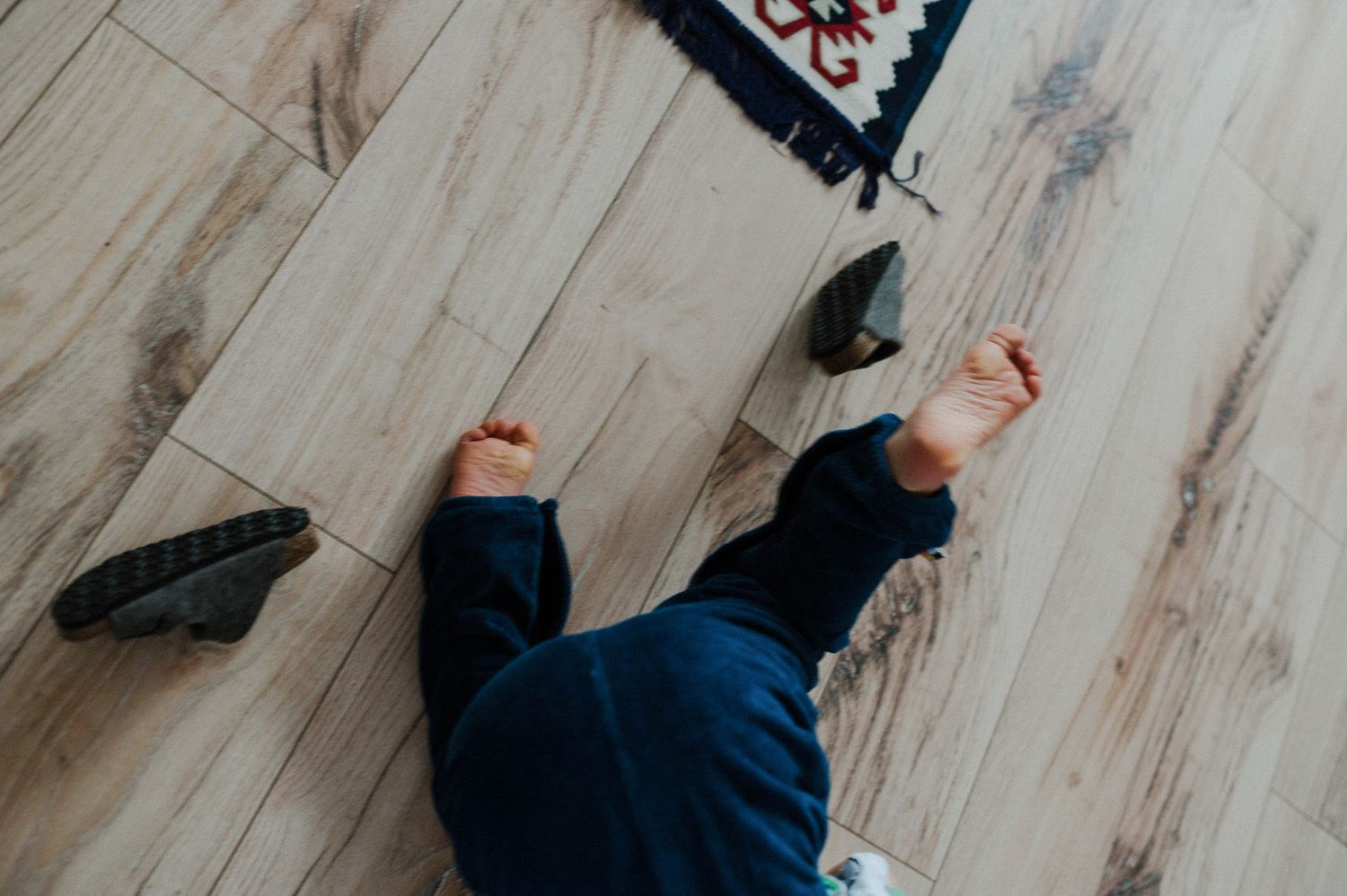 Overhead view of someone lying on a light wooden floor wearing navy pants with black shoes nearby.