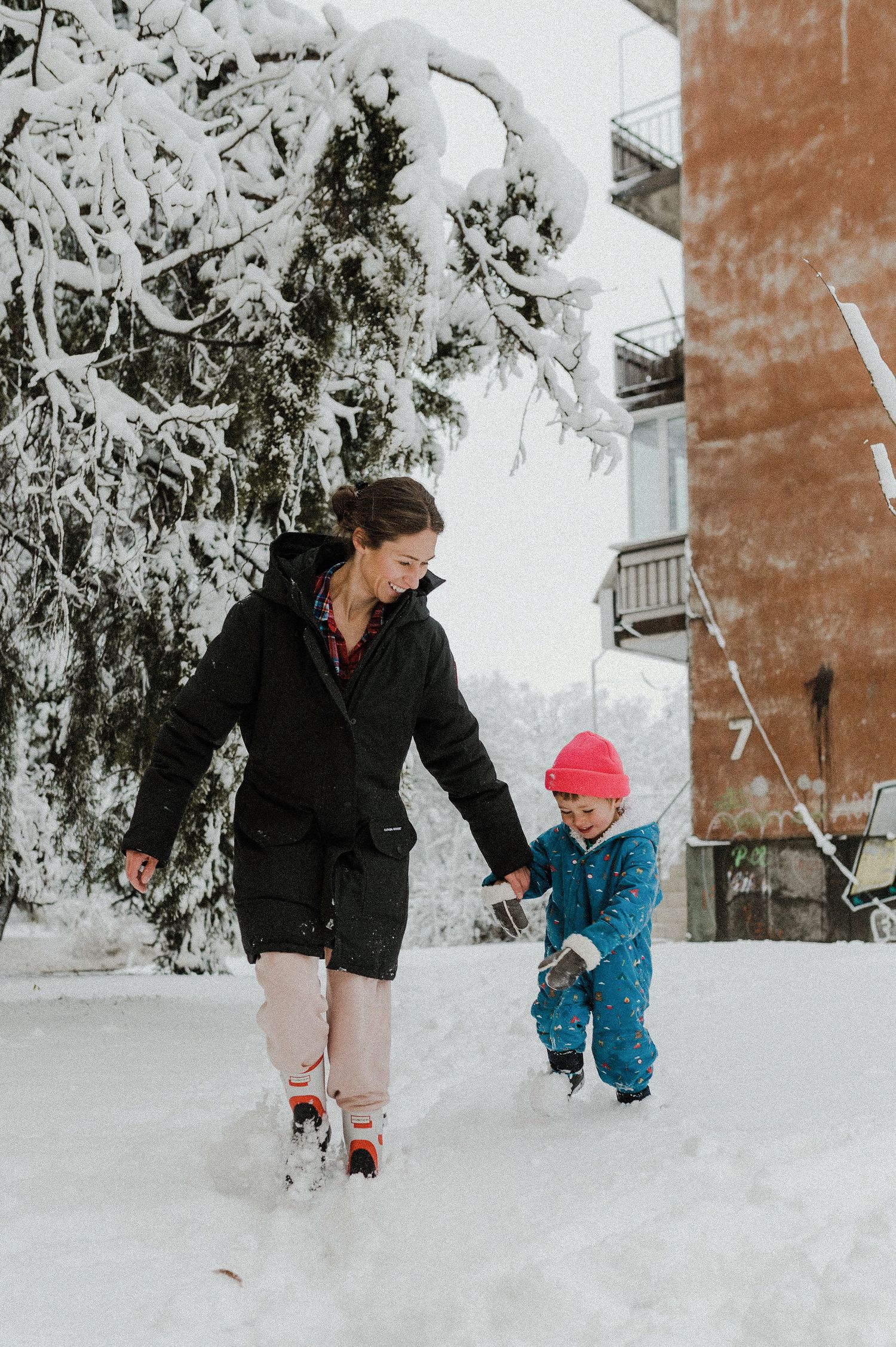 Parent and small child play together in deep winter snow beside snow-covered evergreen trees.