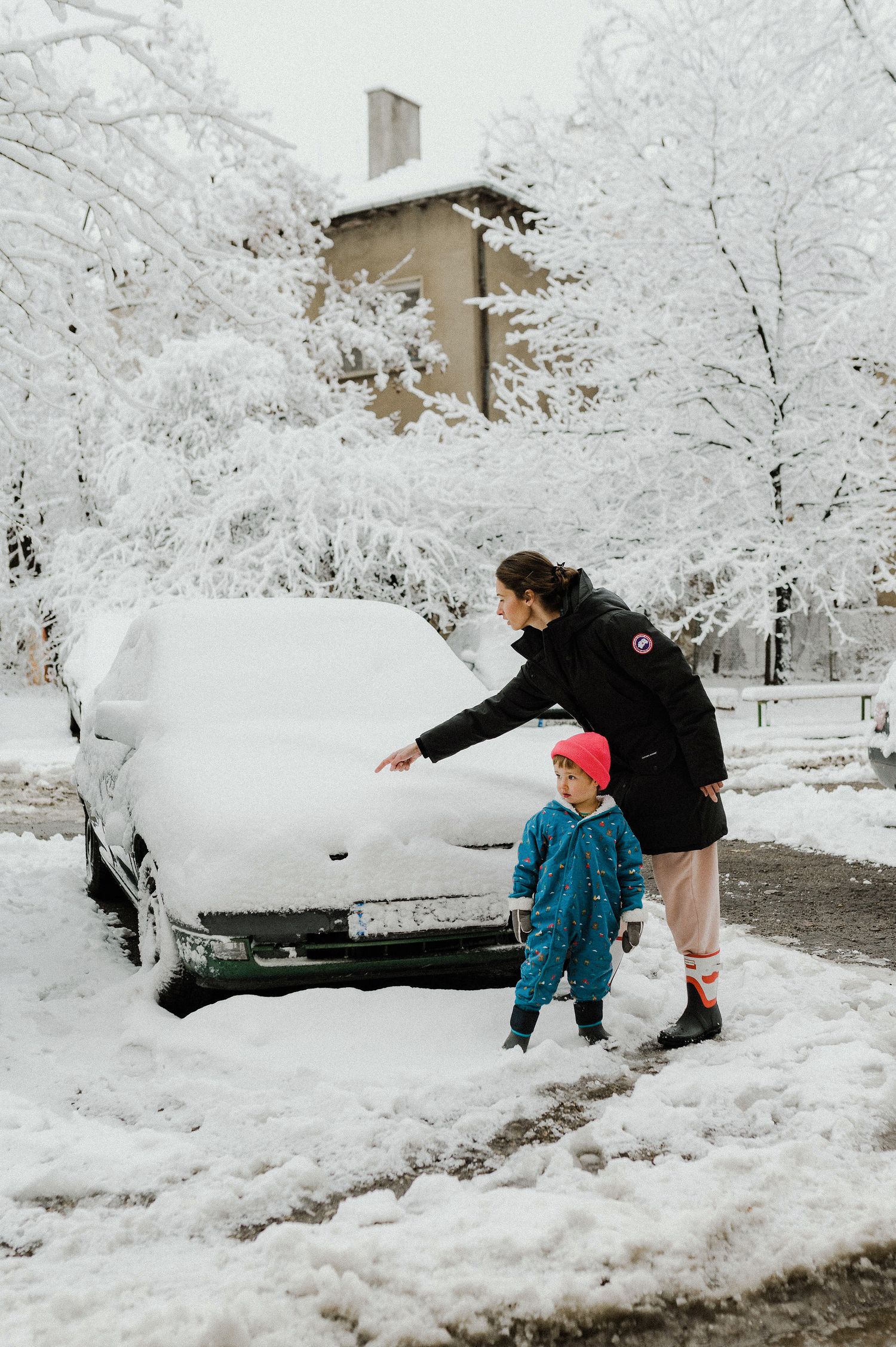 People clearing deep snow off parked cars in a winter storm with snow-covered trees in the background.