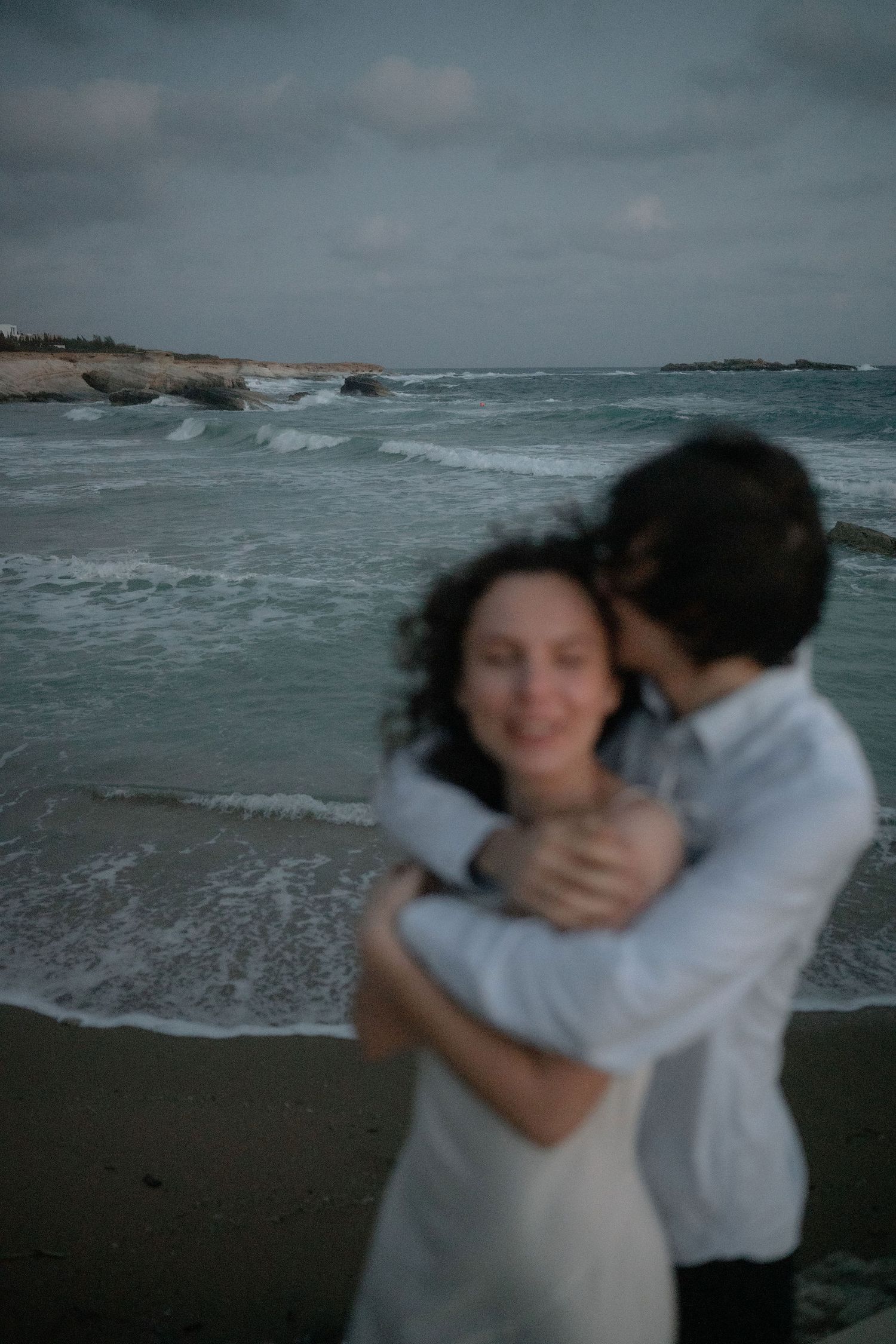 Romantic couple embracing on a rocky beach at dusk, waves crashing behind them in soft focus.