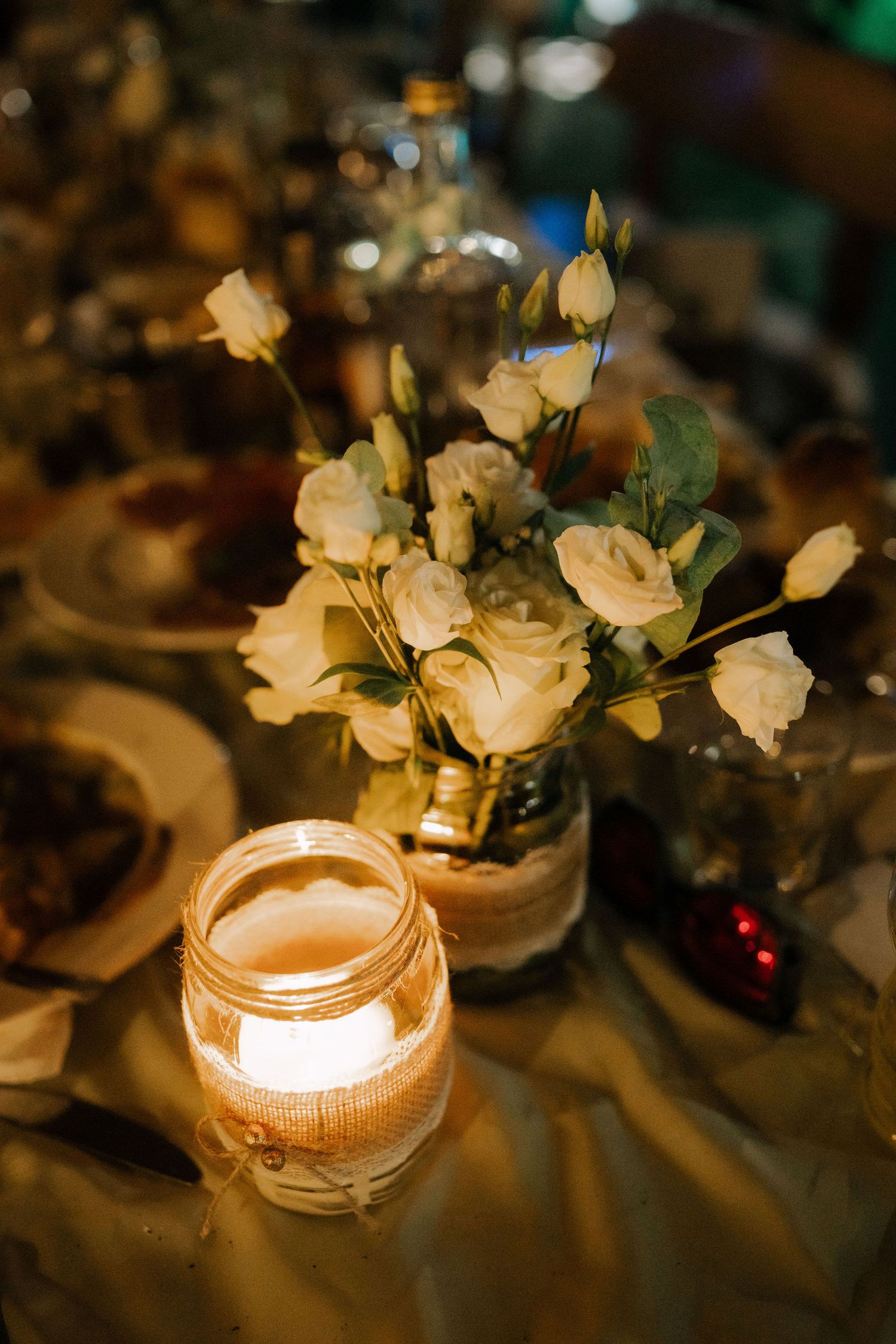 Glowing candles in glass jars adorned with white roses create an elegant table centerpiece in warm lighting.