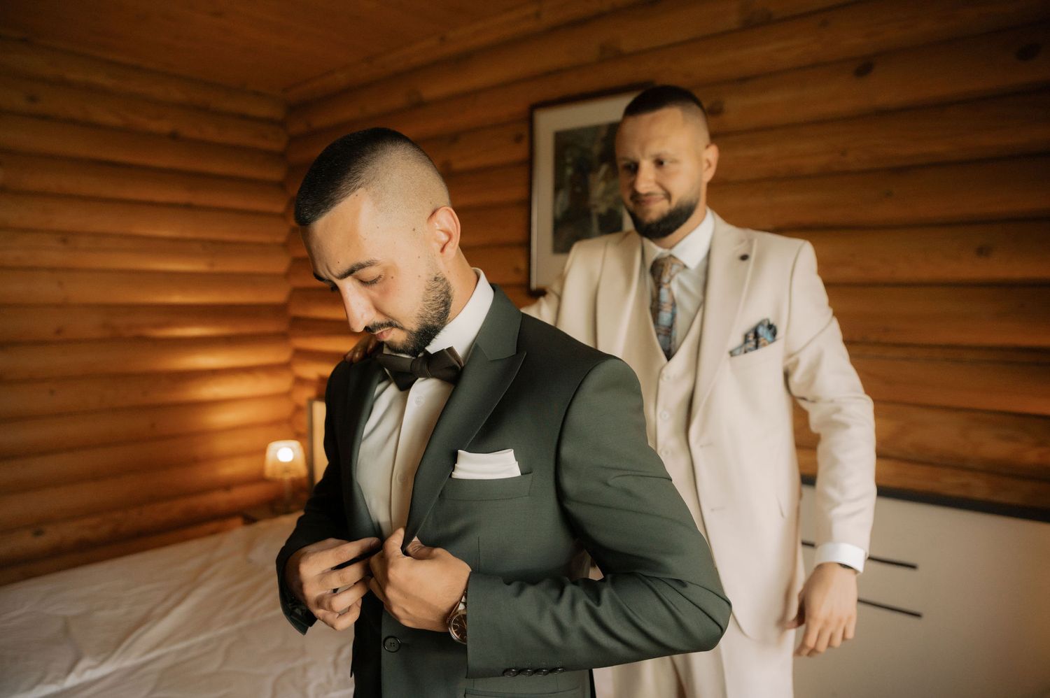 Two men in contrasting suits share a candid moment in a rustic wooden room with warm lighting.