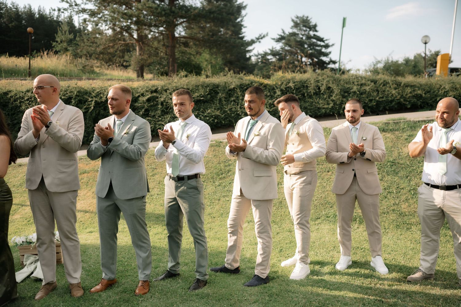 A group of groomsmen in light colored suits stand in a row on grass during an outdoor wedding ceremony.