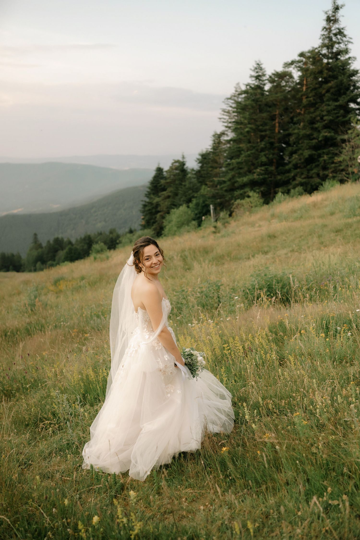 A person in a flowing white wedding dress stands on a mountainside meadow with evergreen trees and misty hills in the background.