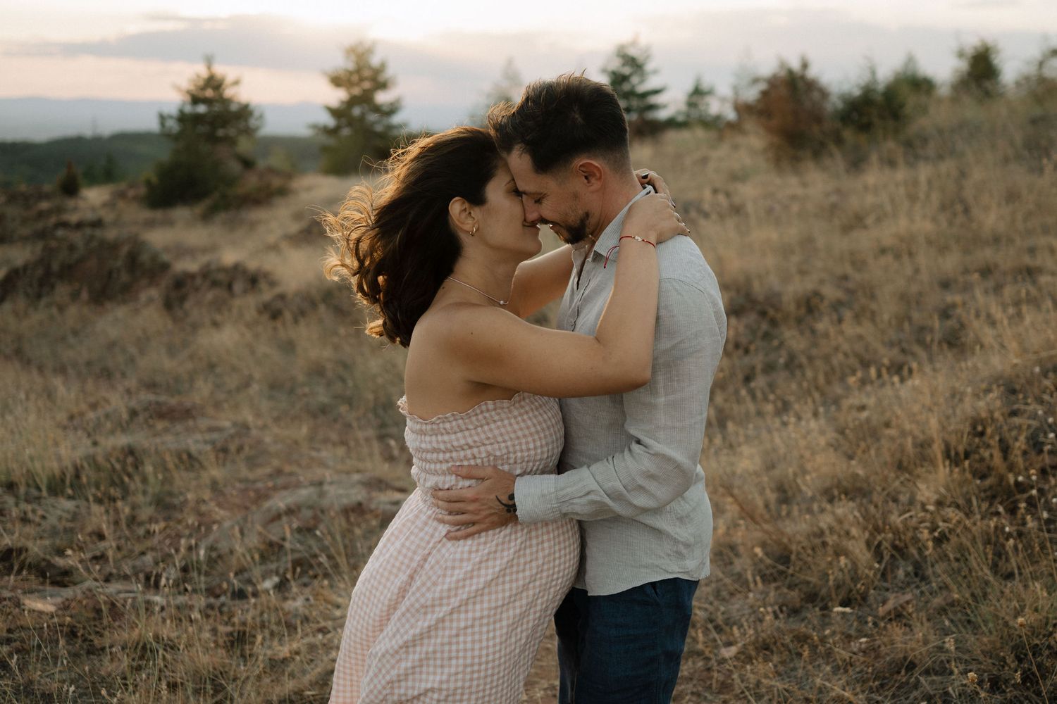 A romantic couple embraces in a golden field at sunset with mountains and evergreen trees in the background.