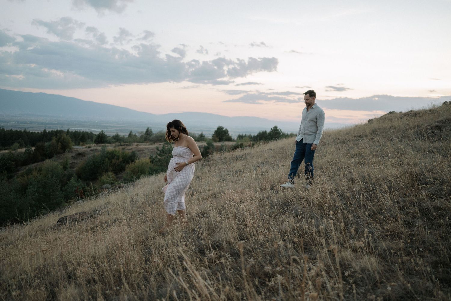 A couple stands together on a golden hillside during sunset with mountains in the distance.