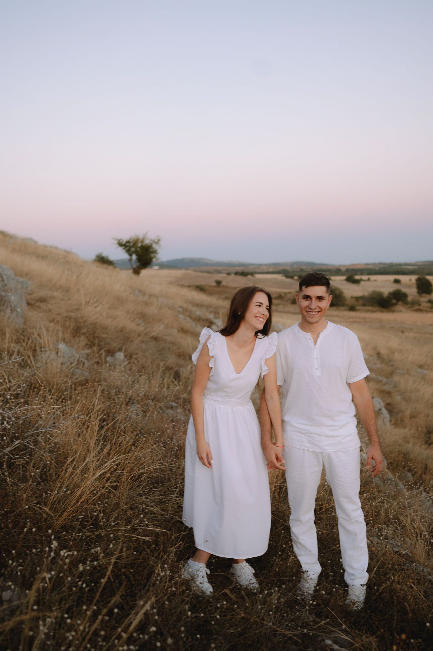 Couple in matching white outfits pose together in a golden field during sunset.