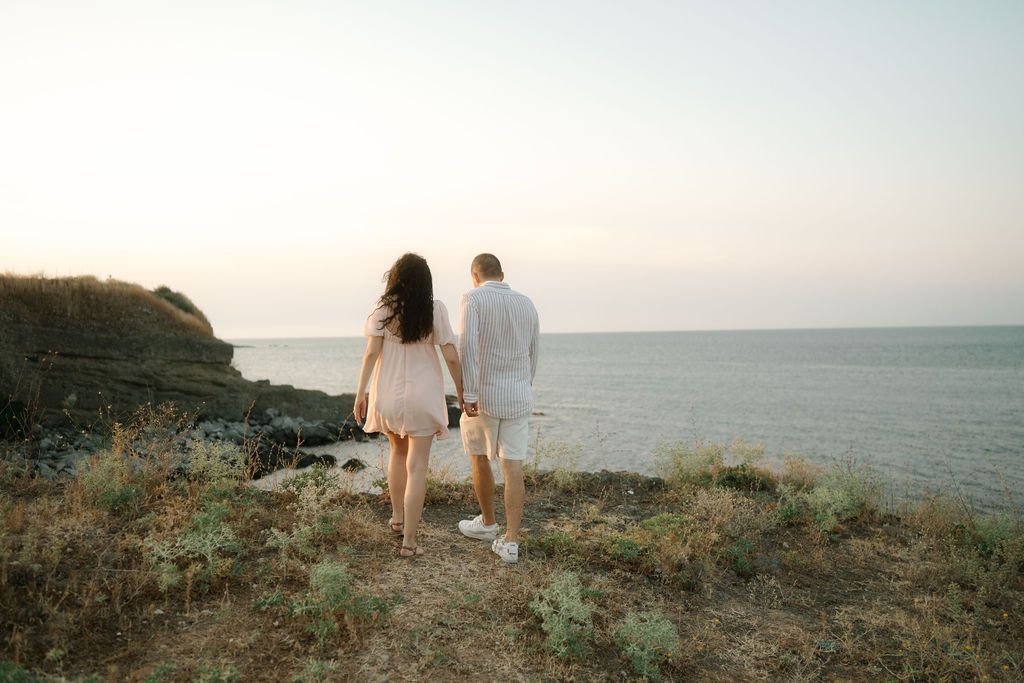 Couple in white outfits walking along a scenic coastal cliff during golden hour.