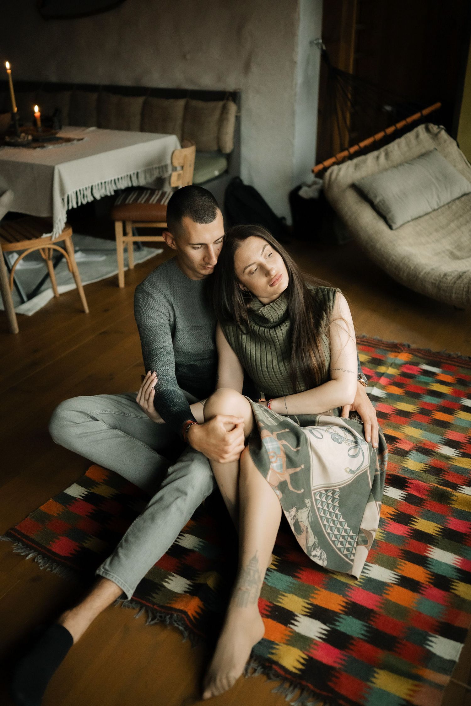A couple sits intimately on a colorful patterned rug in a cozy living room with dim lighting and minimal decor.