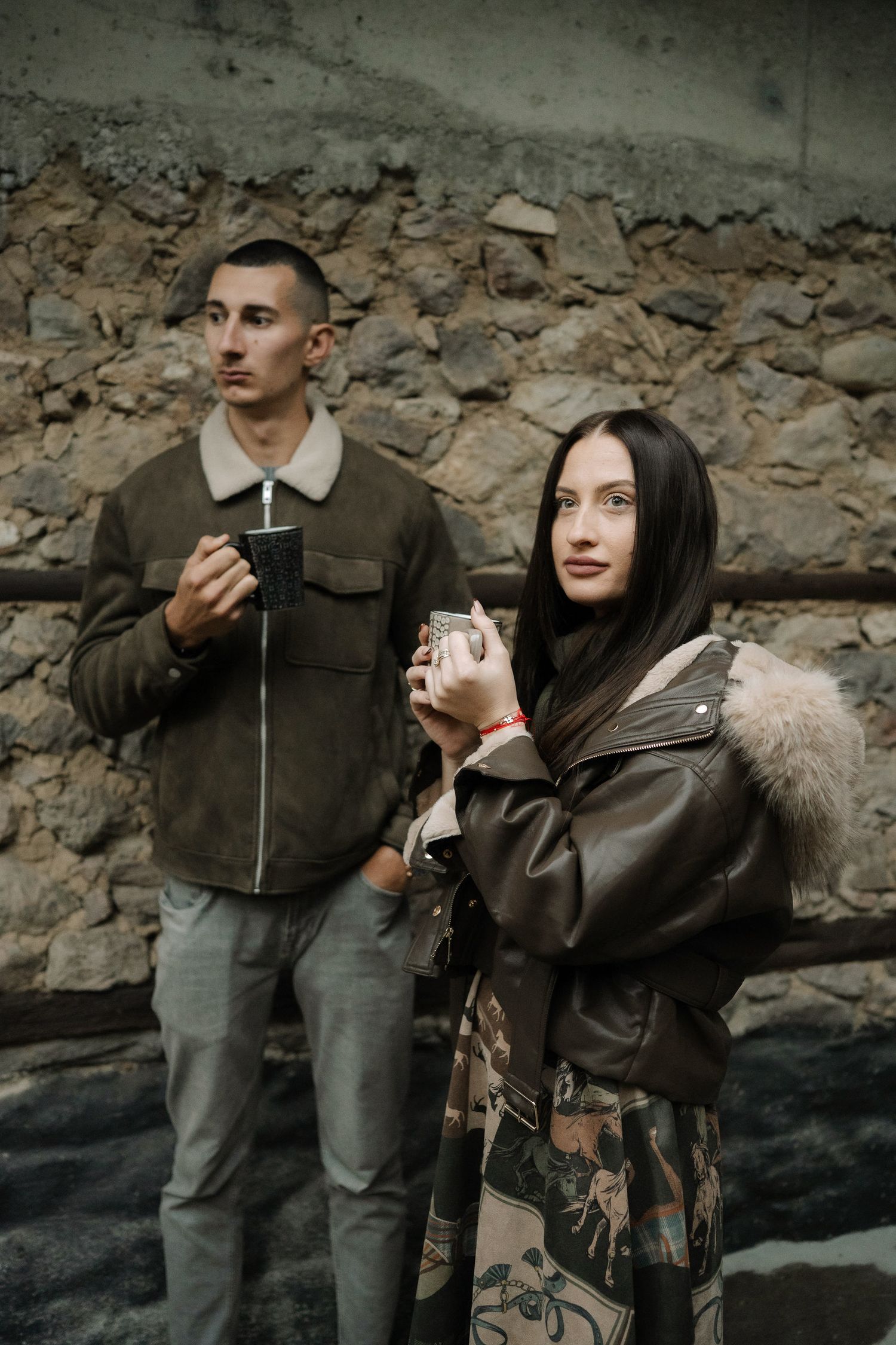 Couple in casual attire sharing warm drinks against a stone wall background.