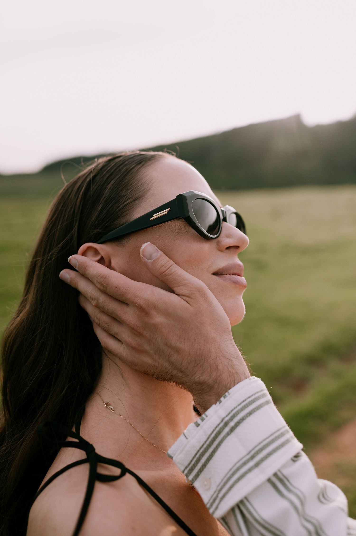 Person wearing sunglasses tilts head back while hands gently touch their face in a serene outdoor setting at sunset.