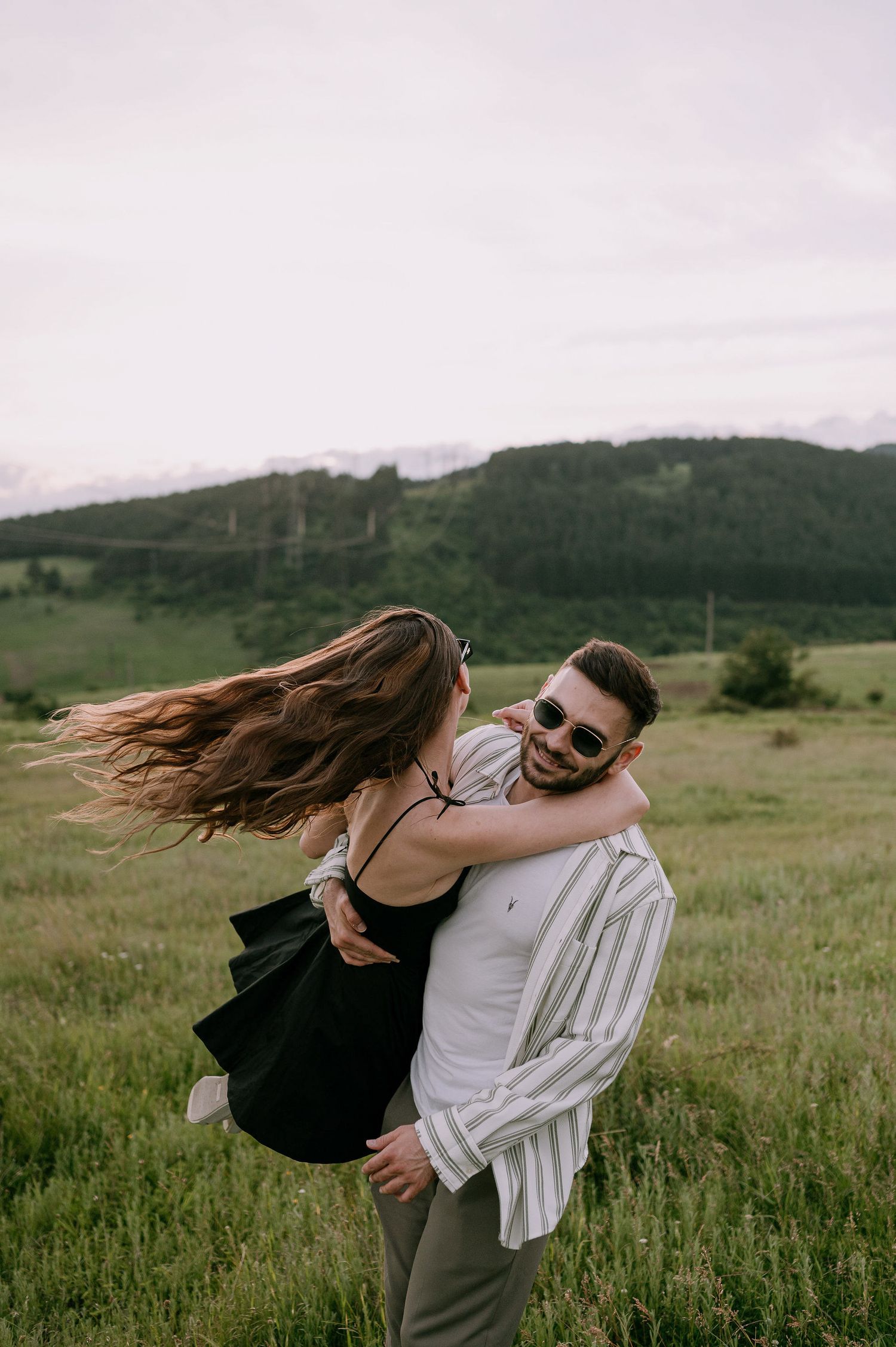 A couple embraces playfully in a grassy meadow with rolling hills in the background on a cloudy day.