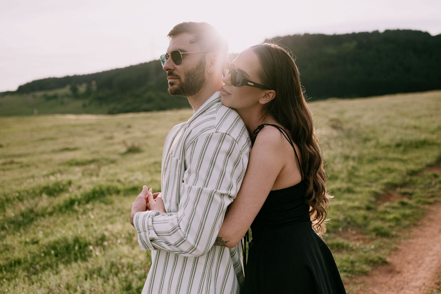 A couple in casual attire walks along a scenic dirt path through rolling green hills during golden hour.