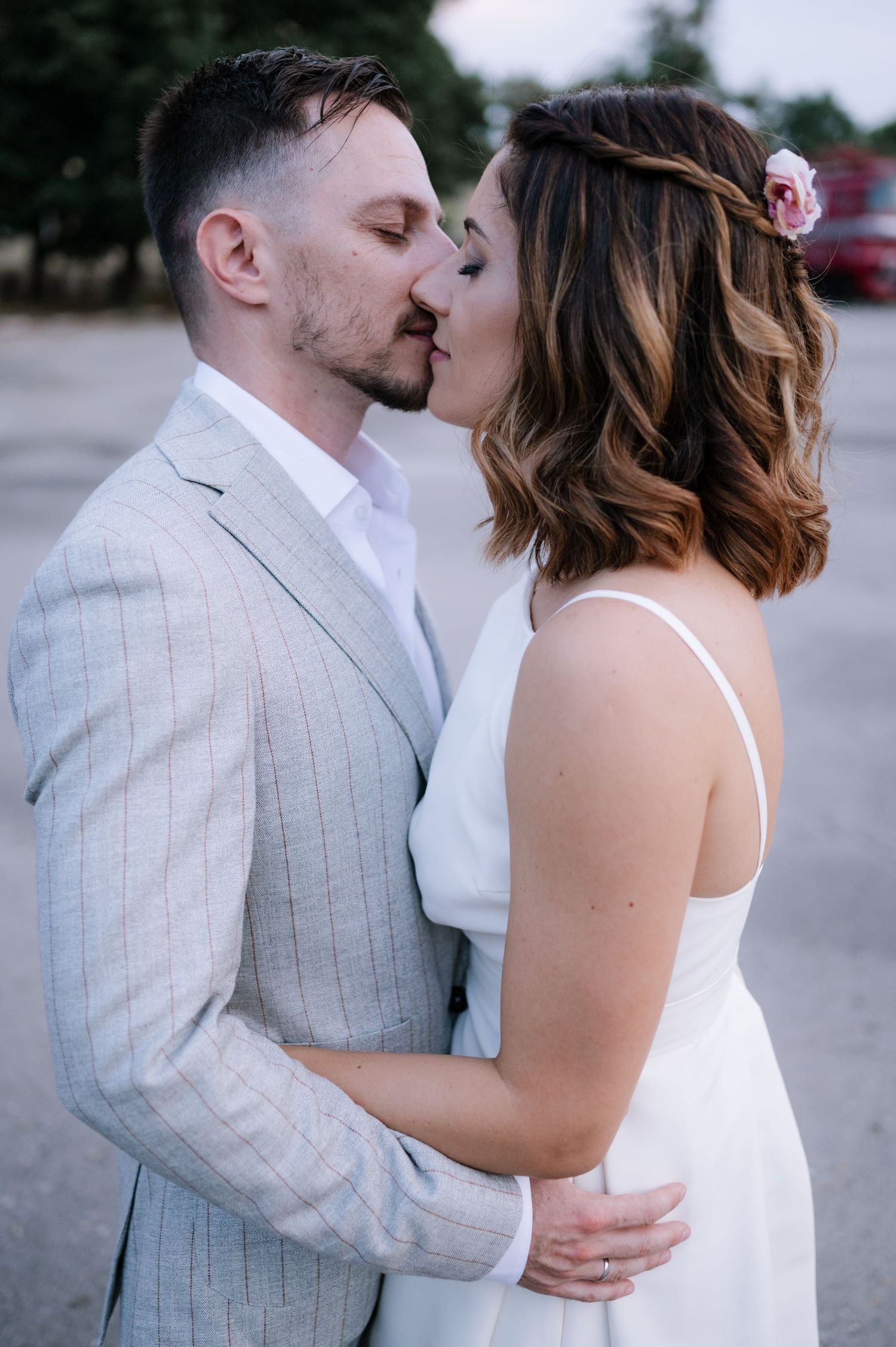 Romantic couple shares tender kisses and embraces while wearing white and light gray formal attire outdoors.