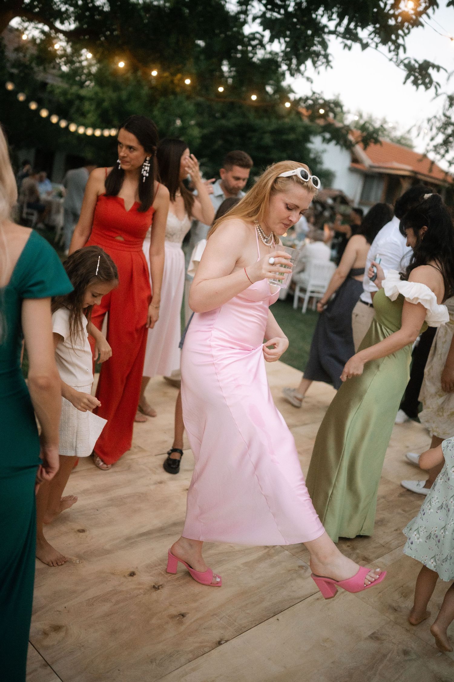Wedding guests dance together outdoors at dusk under string lights on a rustic dance floor during a summer celebration.