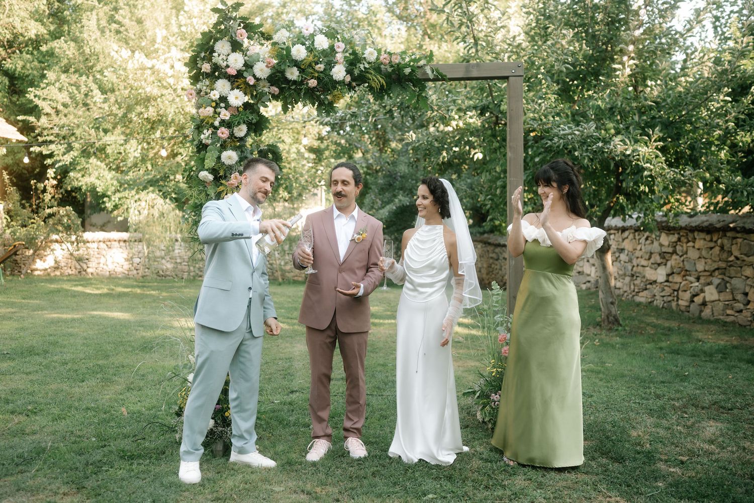 A group of wedding guests and wedding party members stand together in a lush garden setting during a summer celebration.