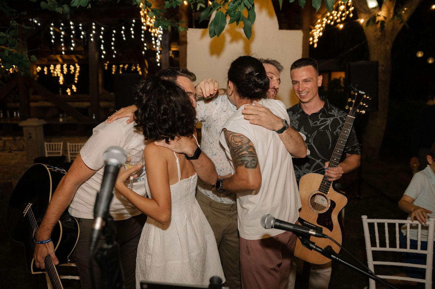 Guests dancing and celebrating at a dimly lit wedding reception with string lights and white chairs in the background.