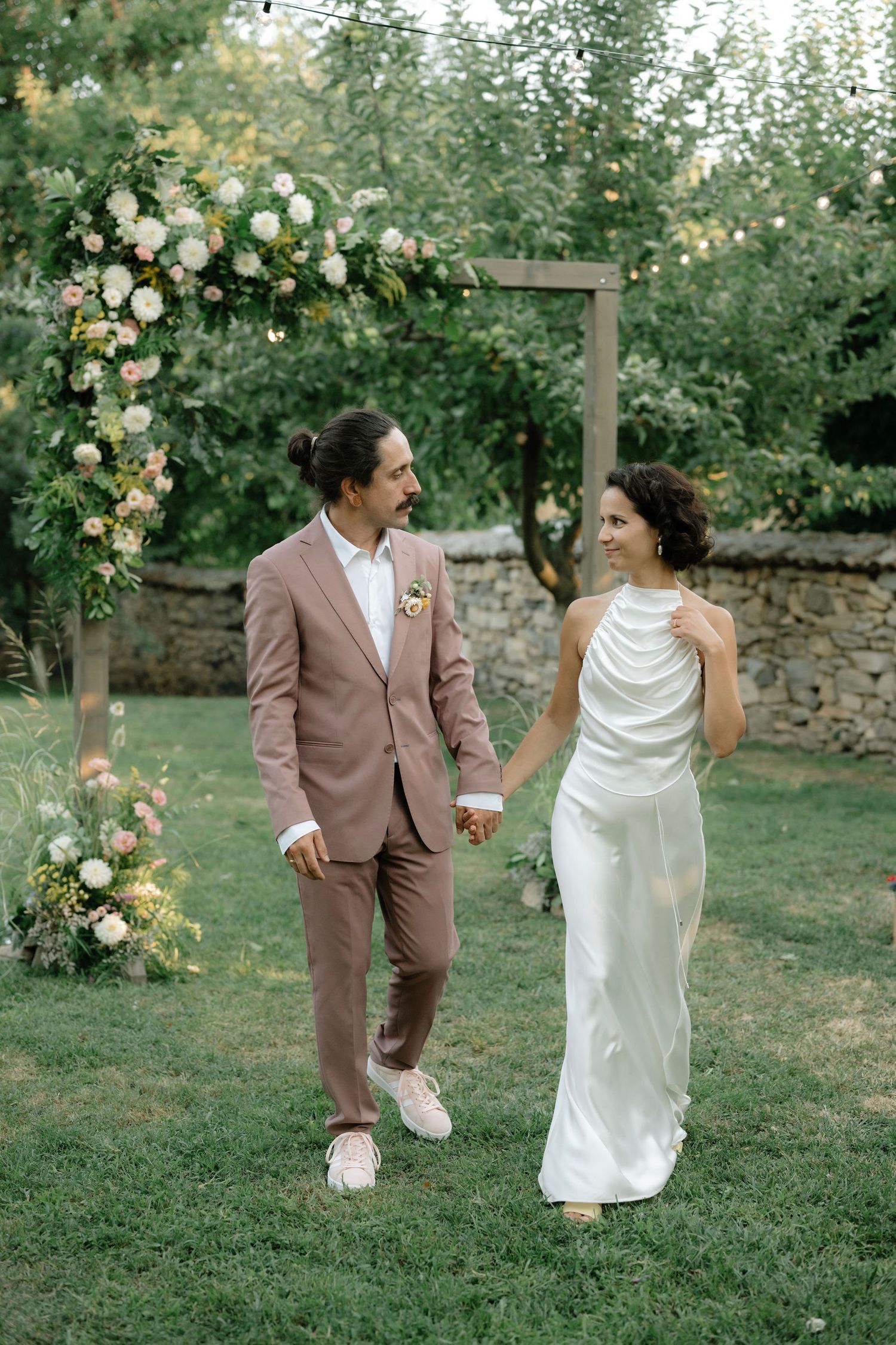 A couple shares a romantic moment under a floral arch in a garden, dressed in elegant wedding attire.