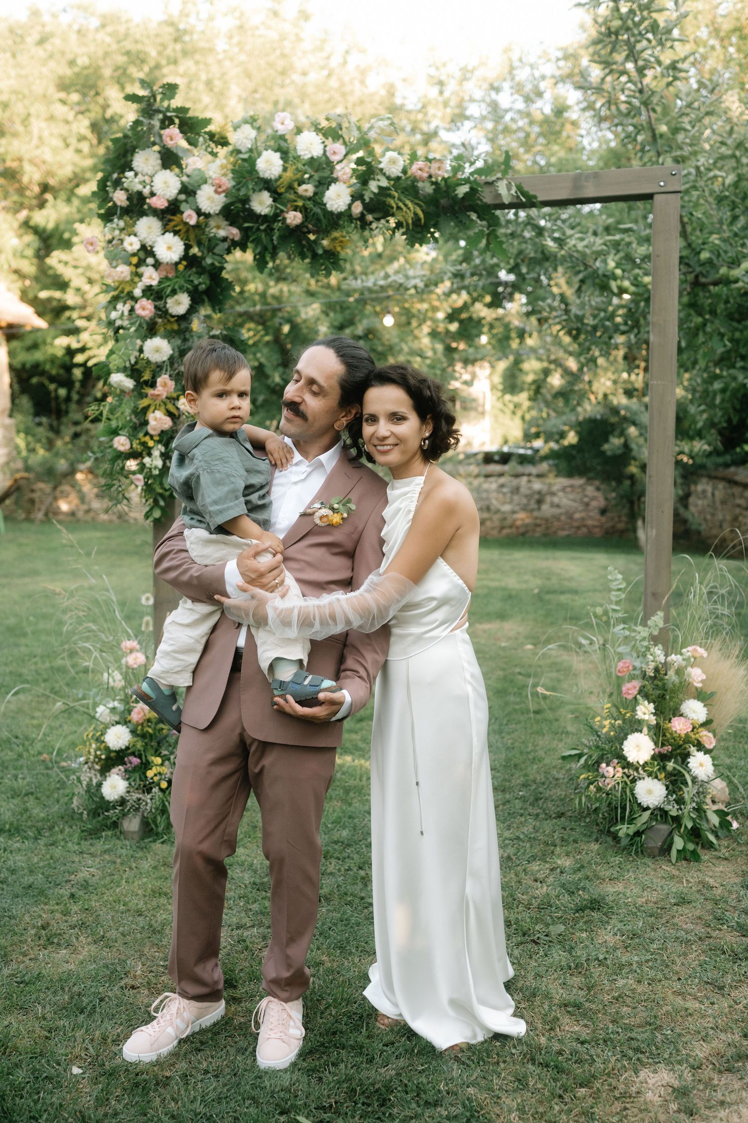 Three people share joyful moments together during an outdoor wedding celebration in a garden setting with white flowers.