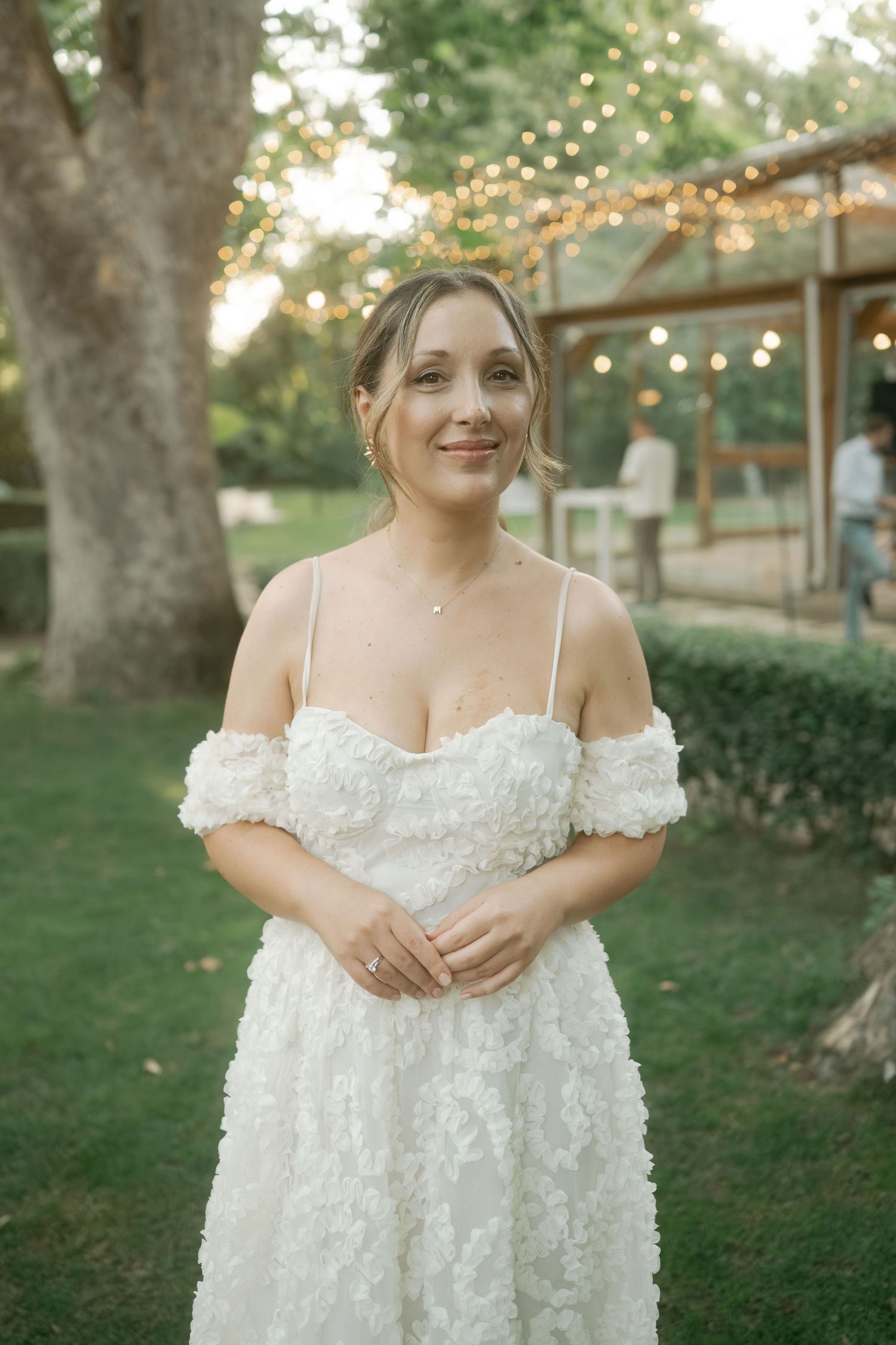 A bride in an off-shoulder white lace wedding dress stands outdoors with string lights and trees in the background.