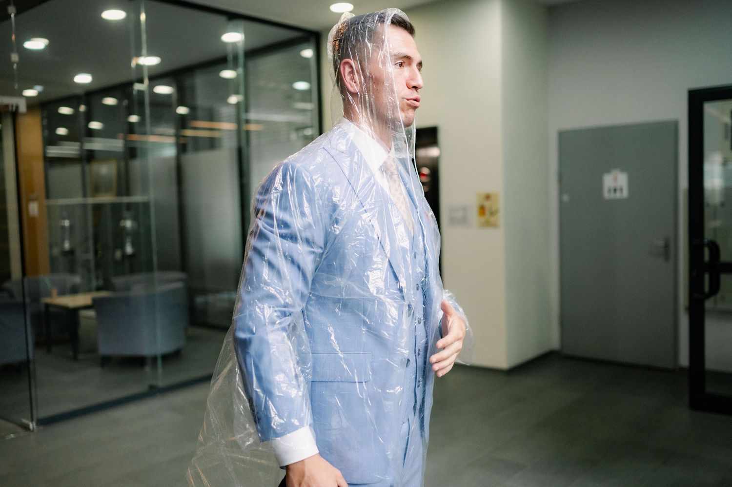 Person in blue shirt walking through modern office hallway with glass walls and overhead lighting.
