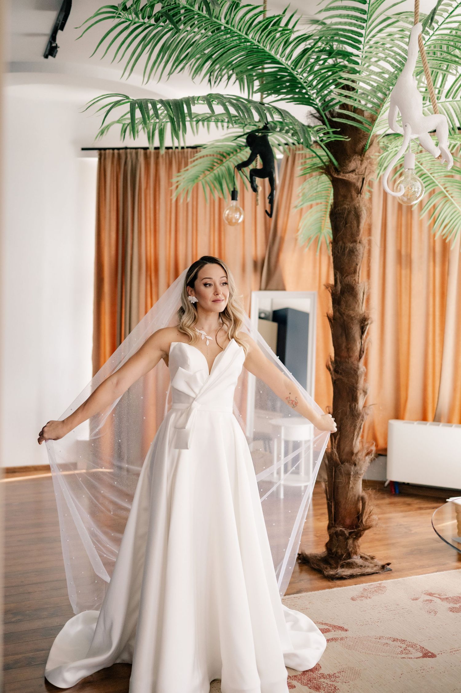 A bride in a flowing white wedding dress poses elegantly near tropical palm decor in an indoor venue.