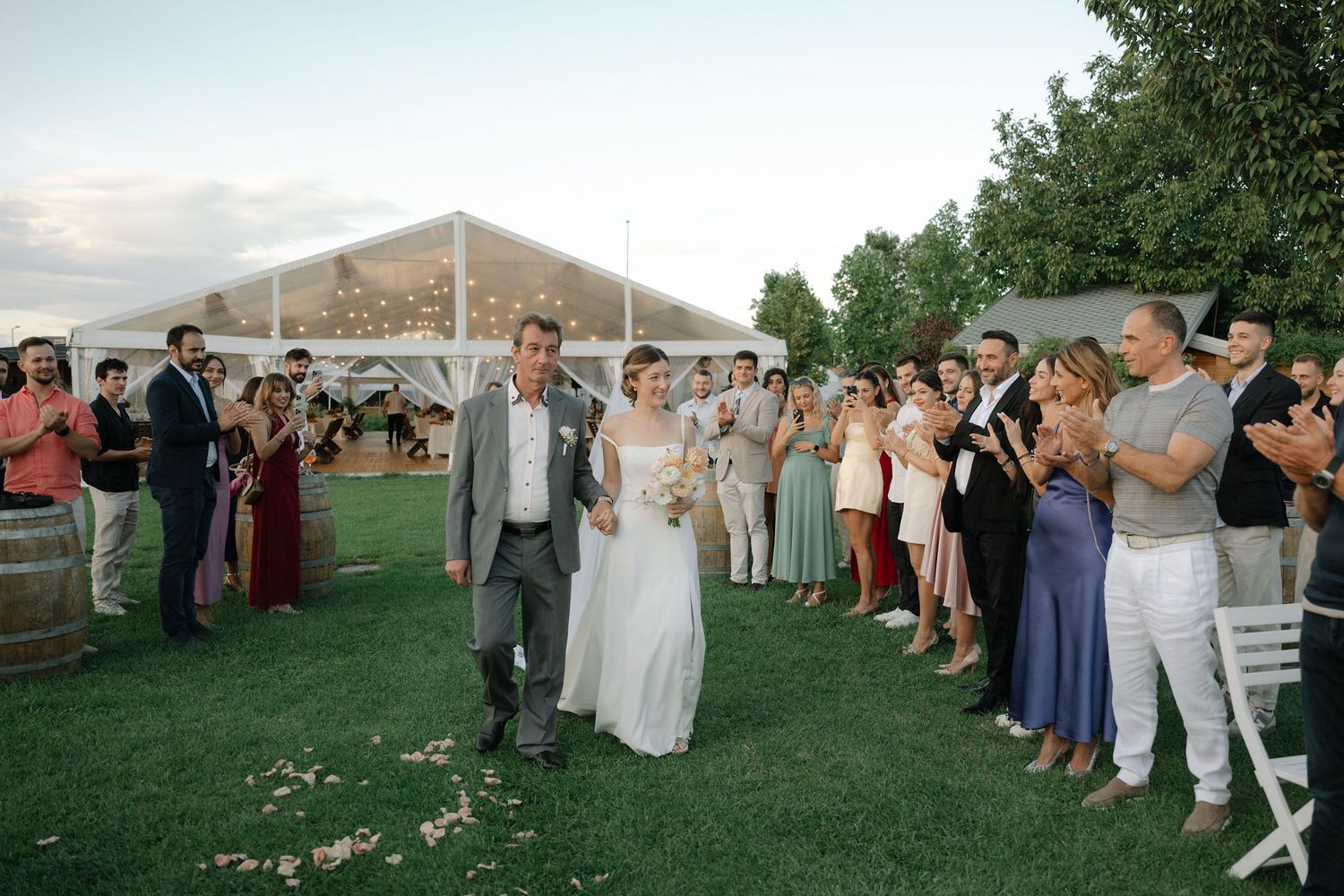 Wedding guests gather outdoors under a tent during sunset for a celebration on a grassy lawn.