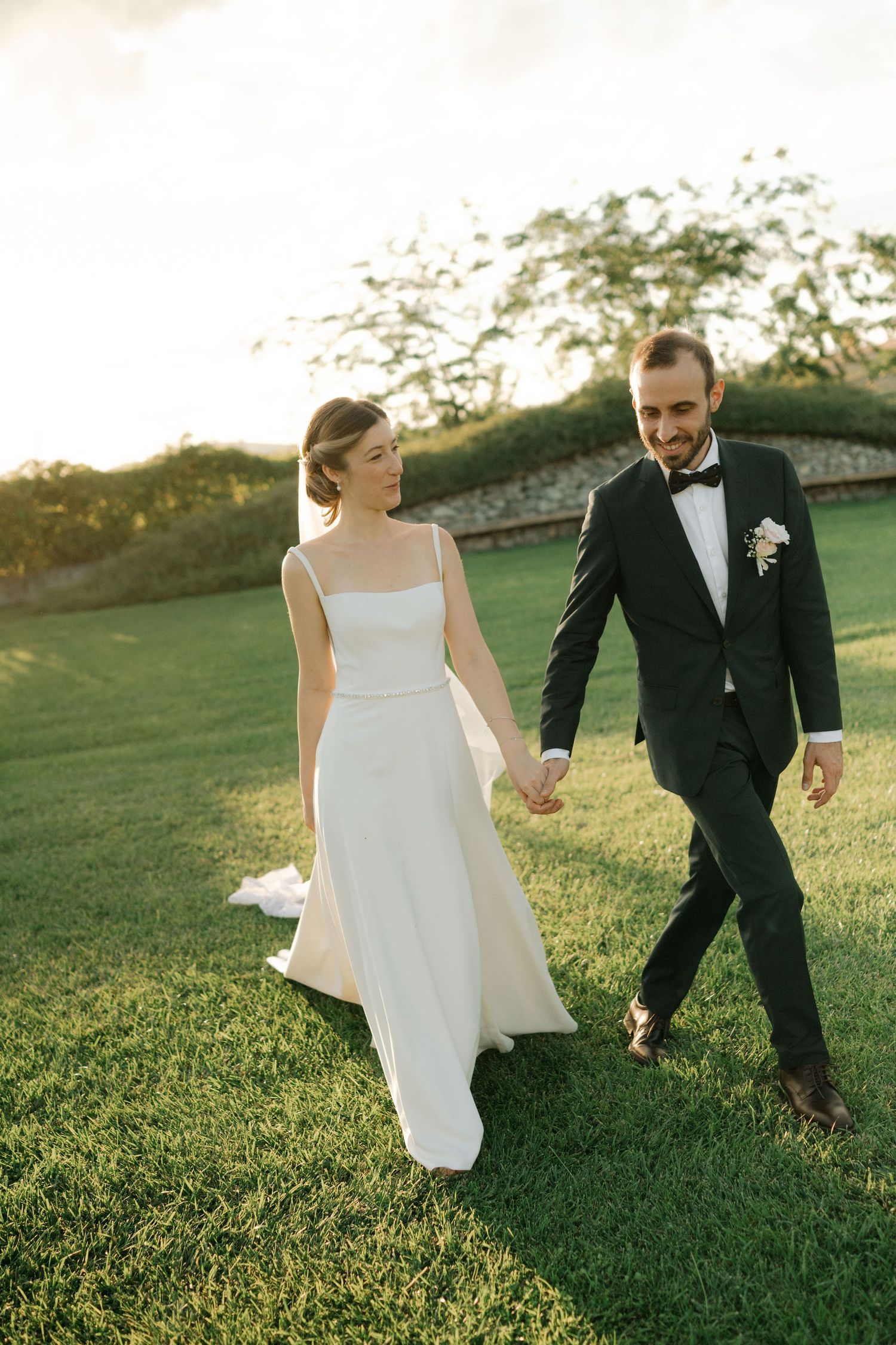 A couple in wedding attire holds hands while walking together on a grassy hill during golden hour.