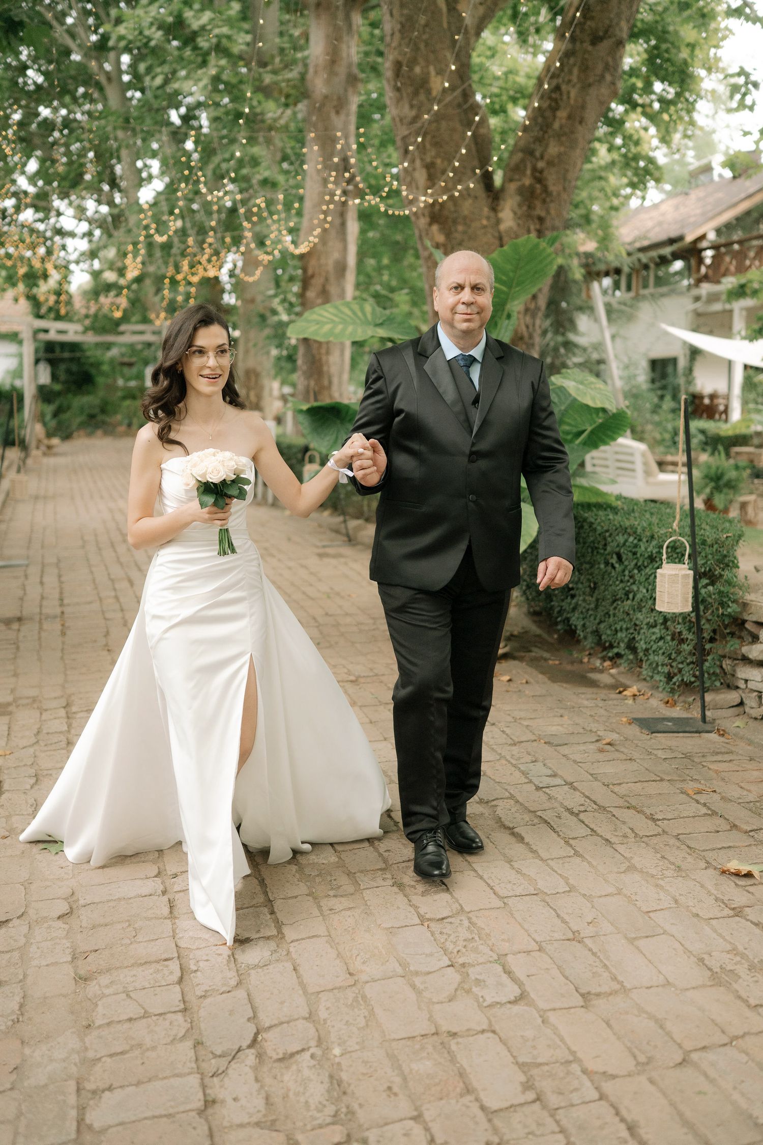 A joyful wedding couple dances playfully on a brick path surrounded by lush green trees.