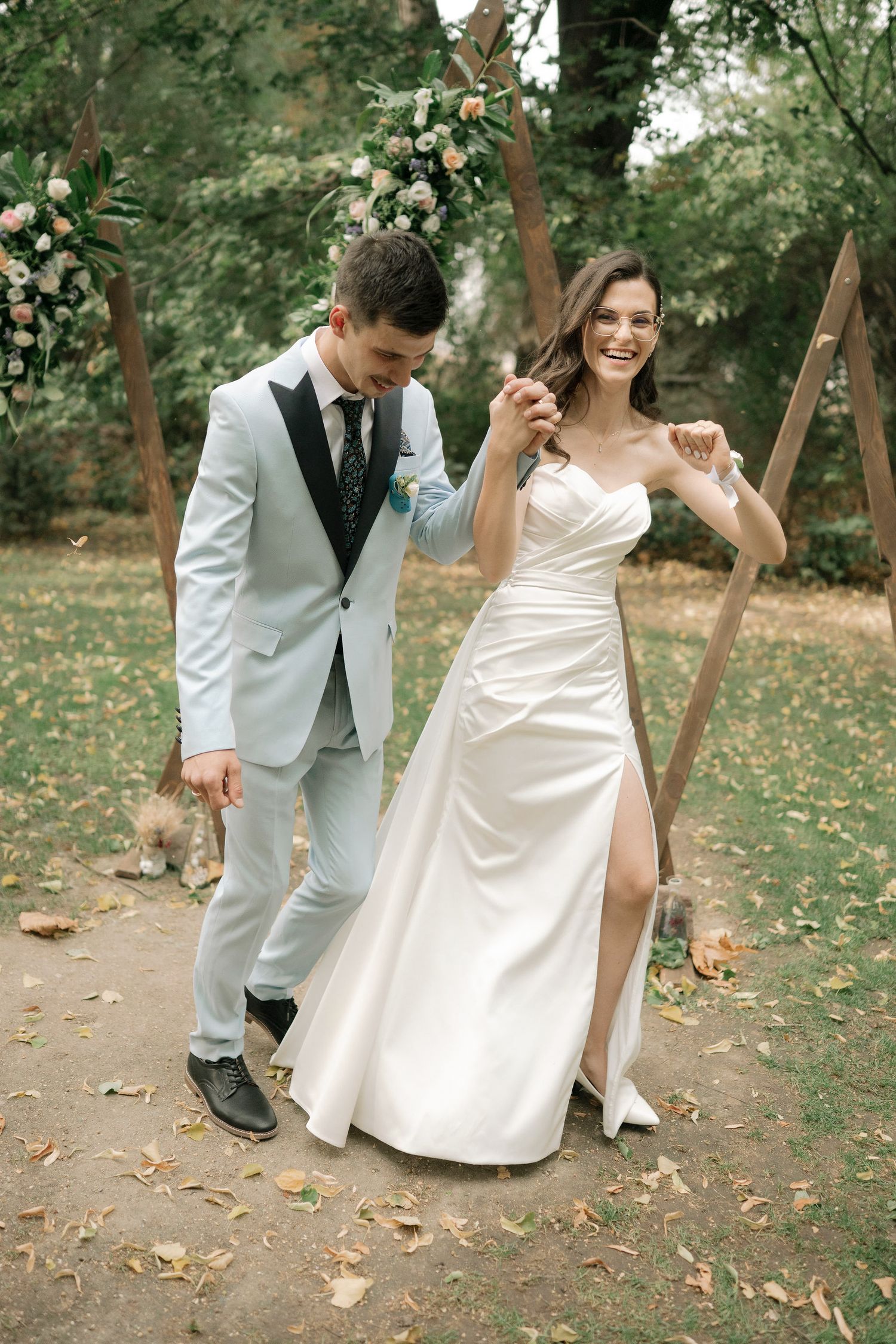 Playful newlyweds dance and twirl together on a leaf-covered forest path during their wedding photoshoot.