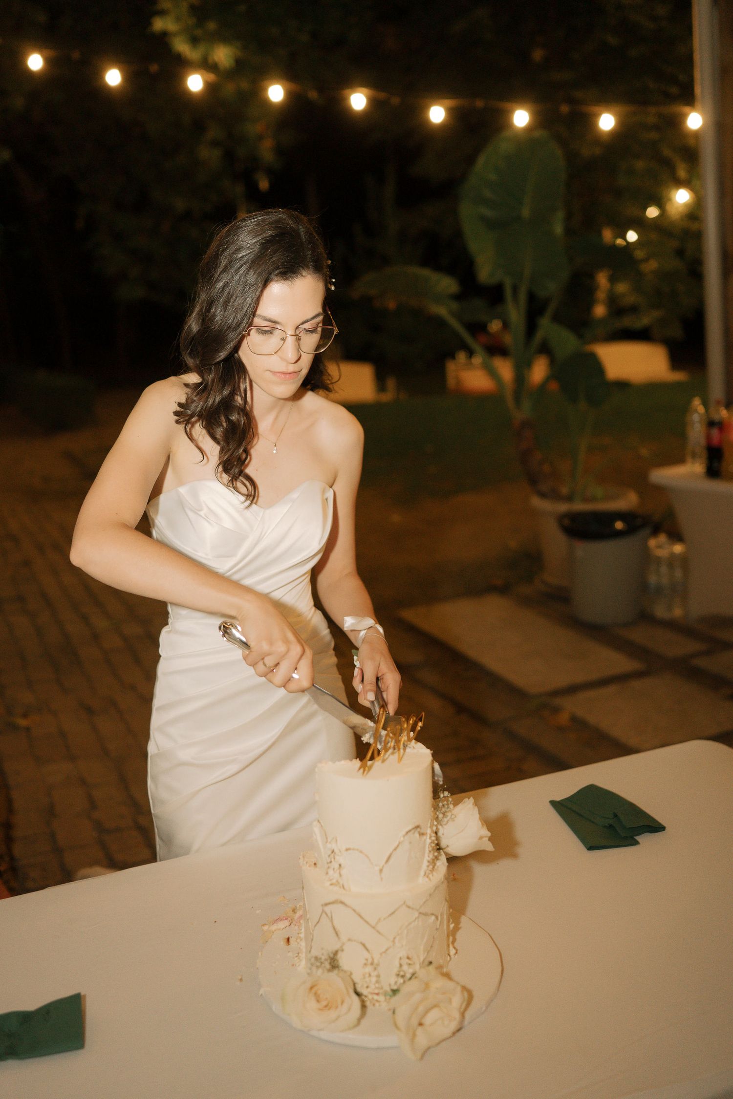 A bride in a white dress cuts into a wedding cake under warm string lights at an evening reception.