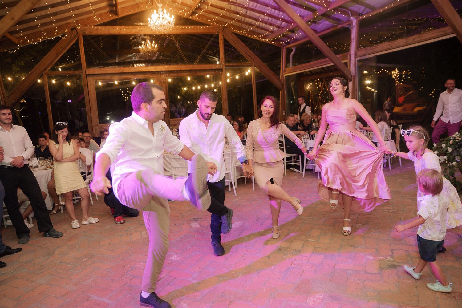 Wedding guests dancing and celebrating on a wooden dance floor under rustic barn rafters with purple mood lighting.