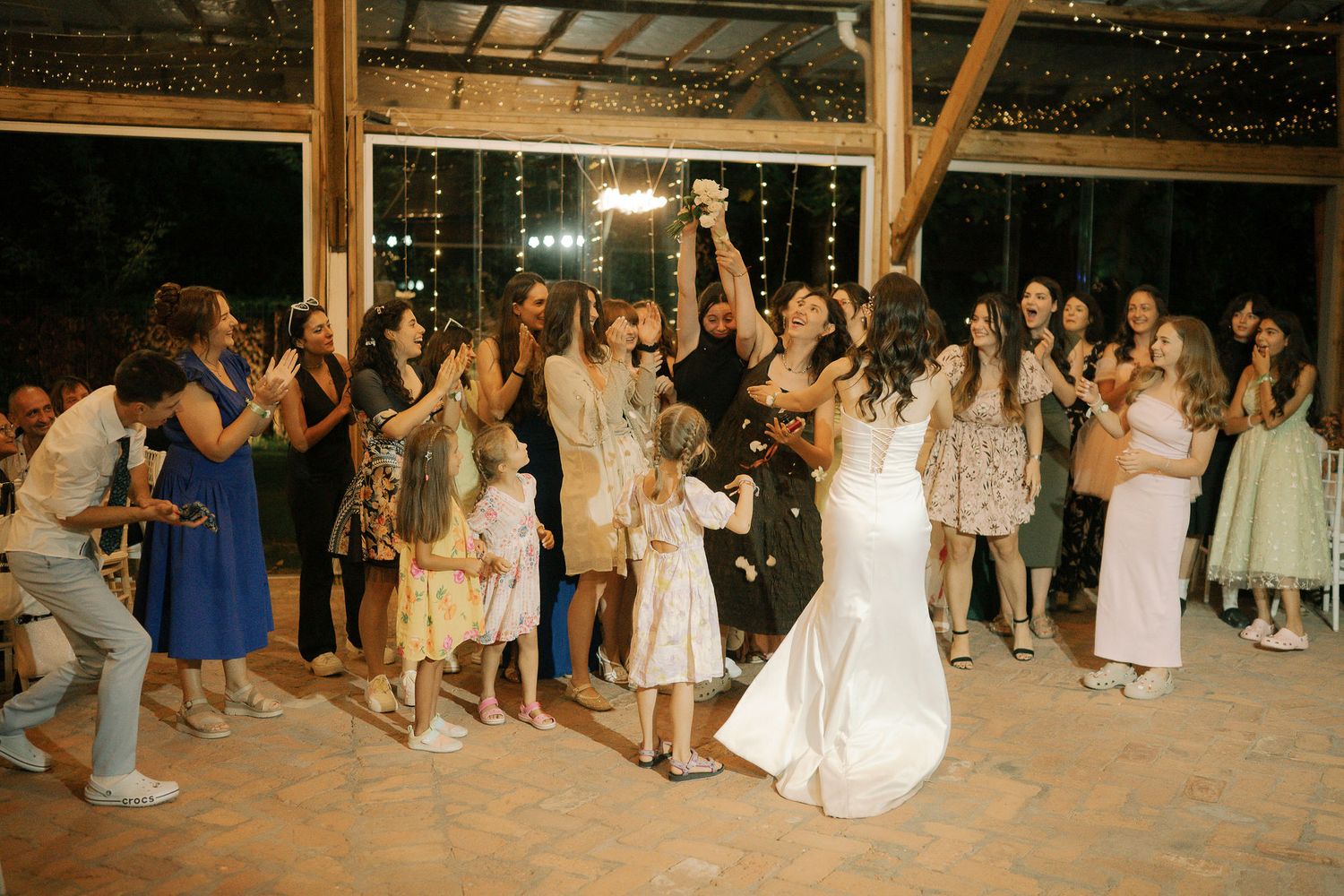 Wedding guests dance in a rustic barn venue decorated with string lights during an evening celebration.