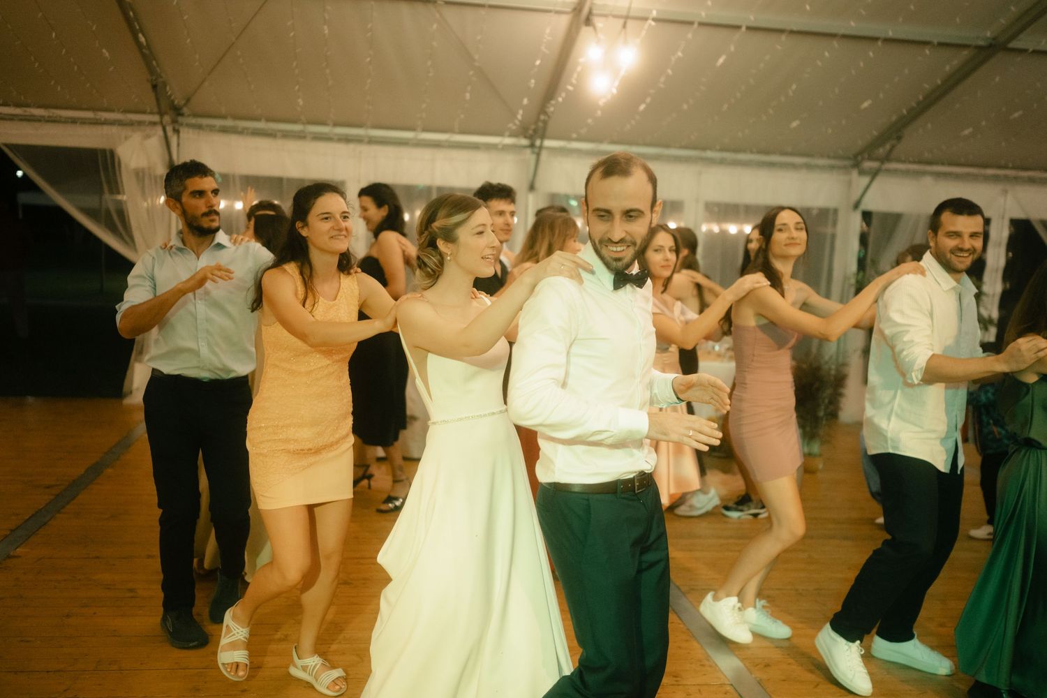 Wedding reception guests dancing in a line on a dance floor under warm lighting at an indoor venue.