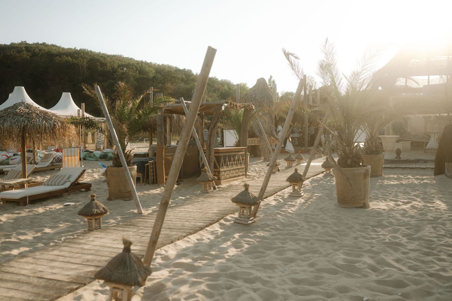 Beach goers enjoy swings and umbrellas at a sandy beachfront bar during golden hour sunset.