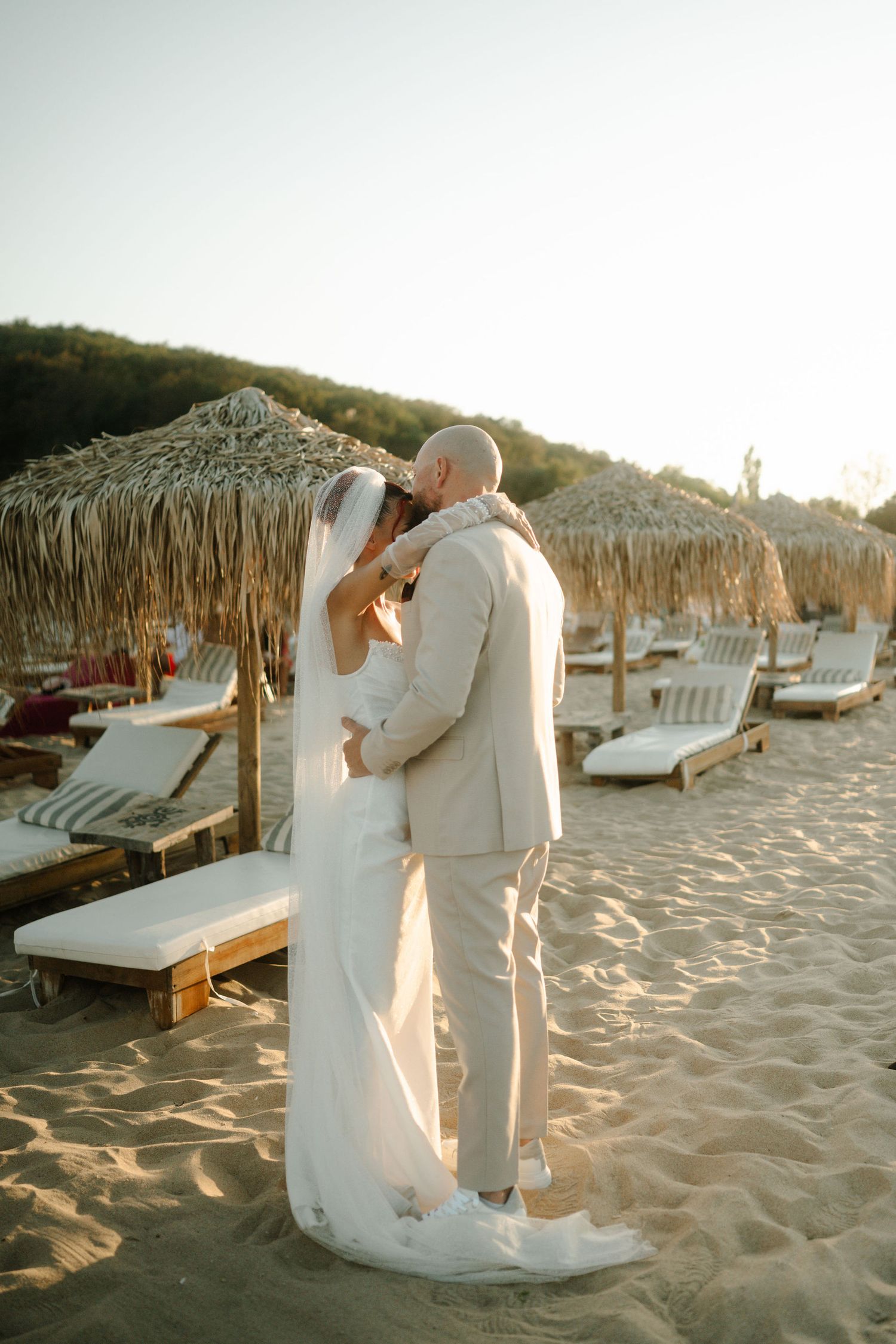 A tender moment between newlyweds on a beach with straw umbrellas and lounge chairs at golden hour.