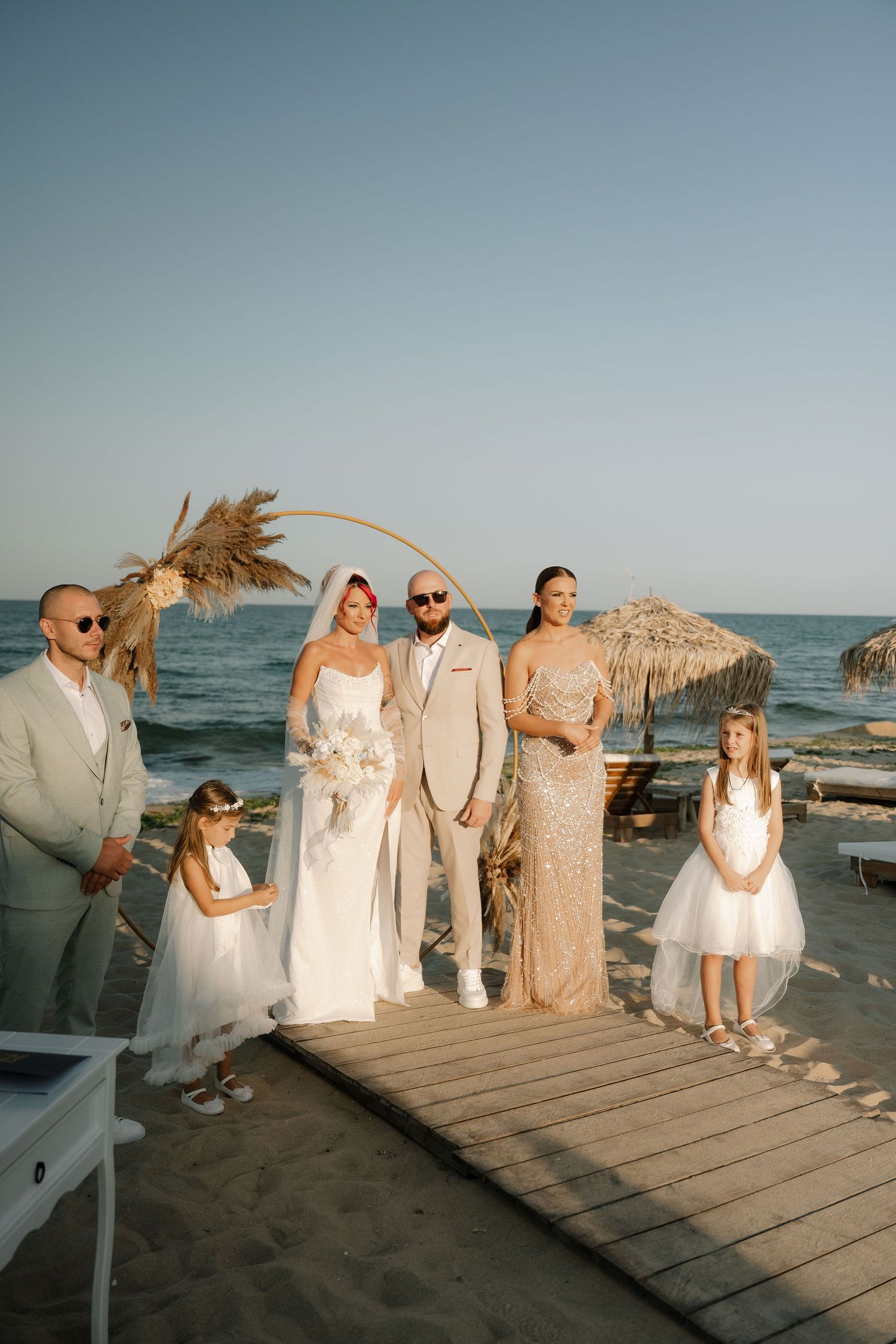 Wedding party in light-colored attire poses on wooden boardwalk at sunset beach with ocean backdrop.
