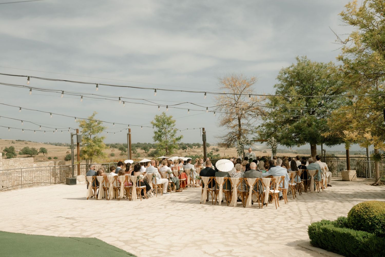 Outdoor wedding ceremony with guests seated on wooden chairs under string lights on a sunny day.