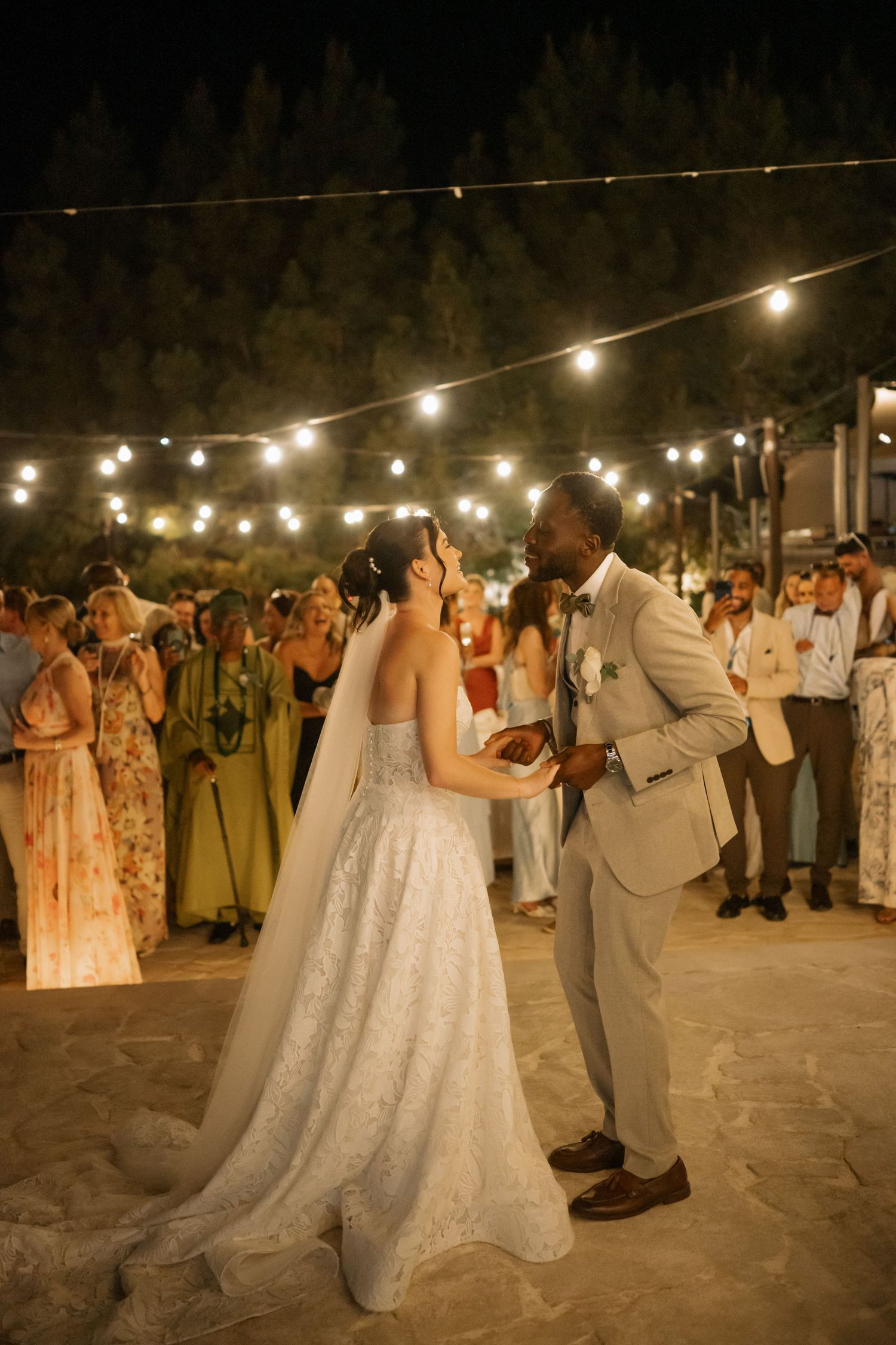 Bride and groom share first dance under string lights at outdoor evening wedding reception with guests watching.