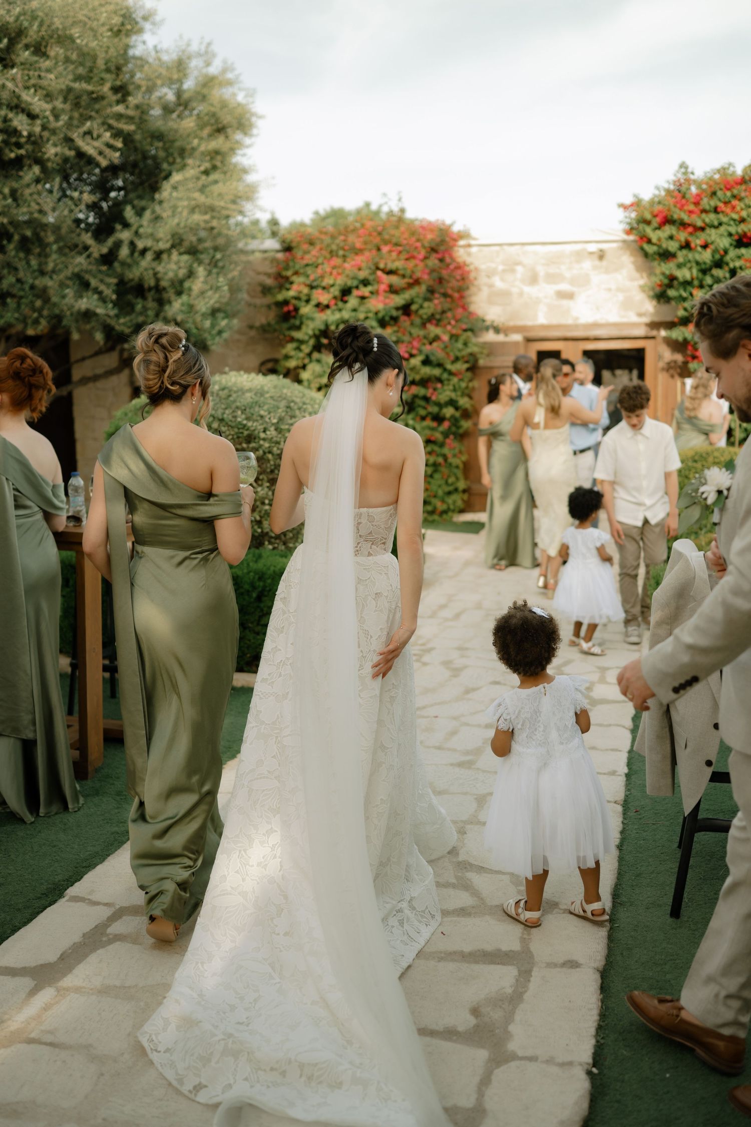 Bride in lace wedding gown and veil walks with bridesmaids in sage green dresses and flower girl at outdoor ceremony.