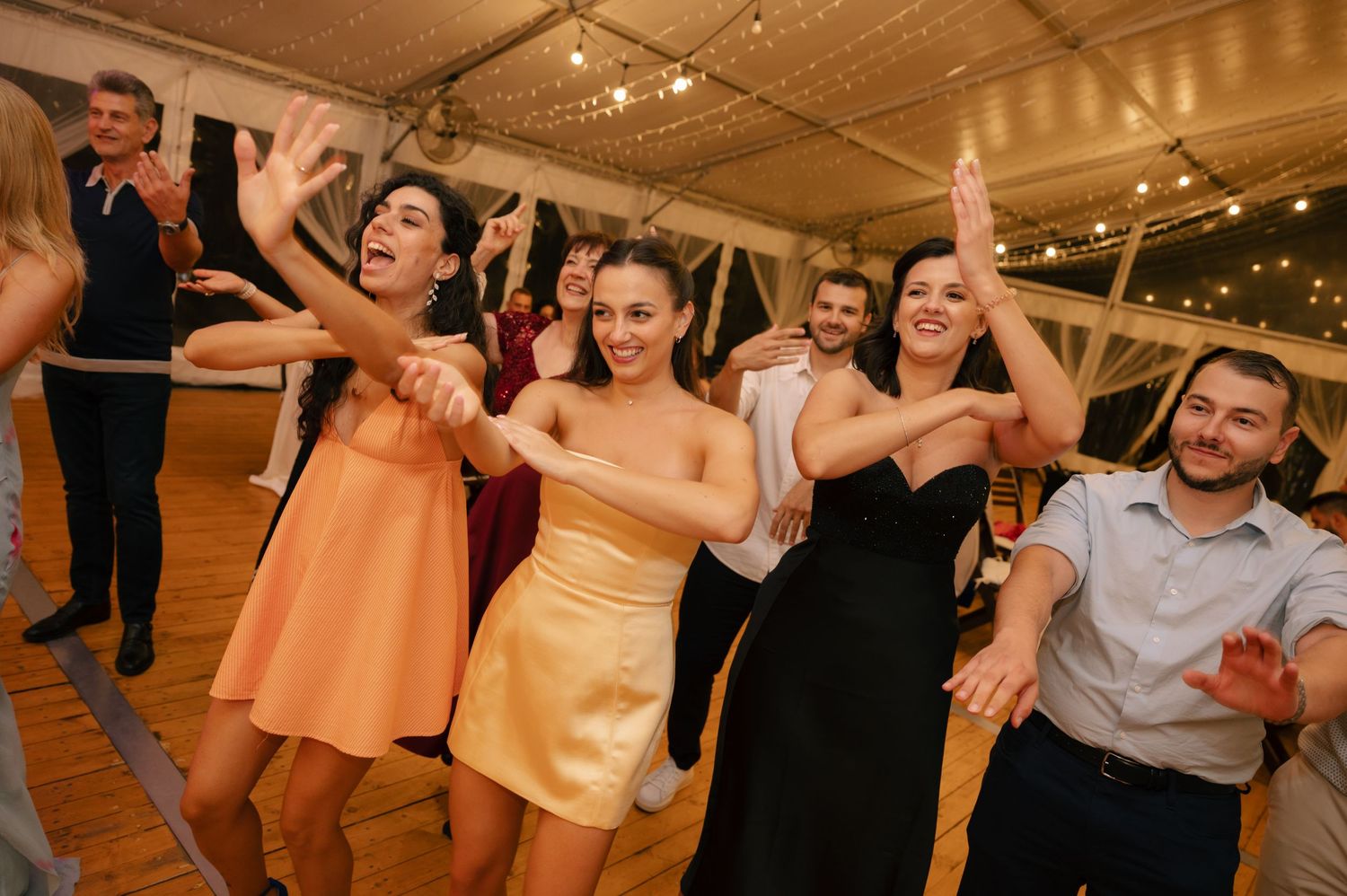 Wedding guests dancing and celebrating together on a dance floor under string lights at an indoor reception venue.