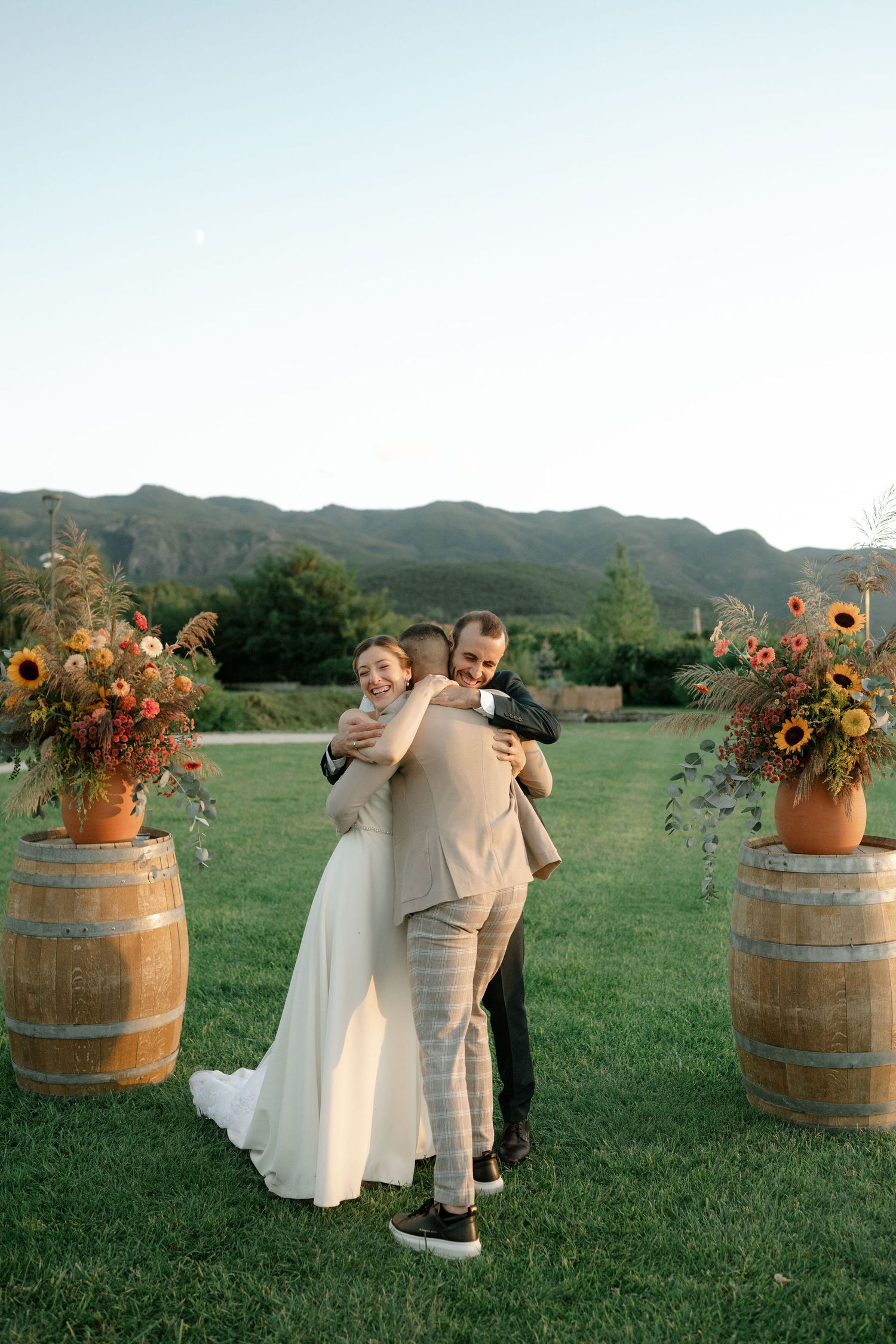 A couple embraces in front of rustic wine barrels decorated with sunflowers on a grassy field with mountains in the background.