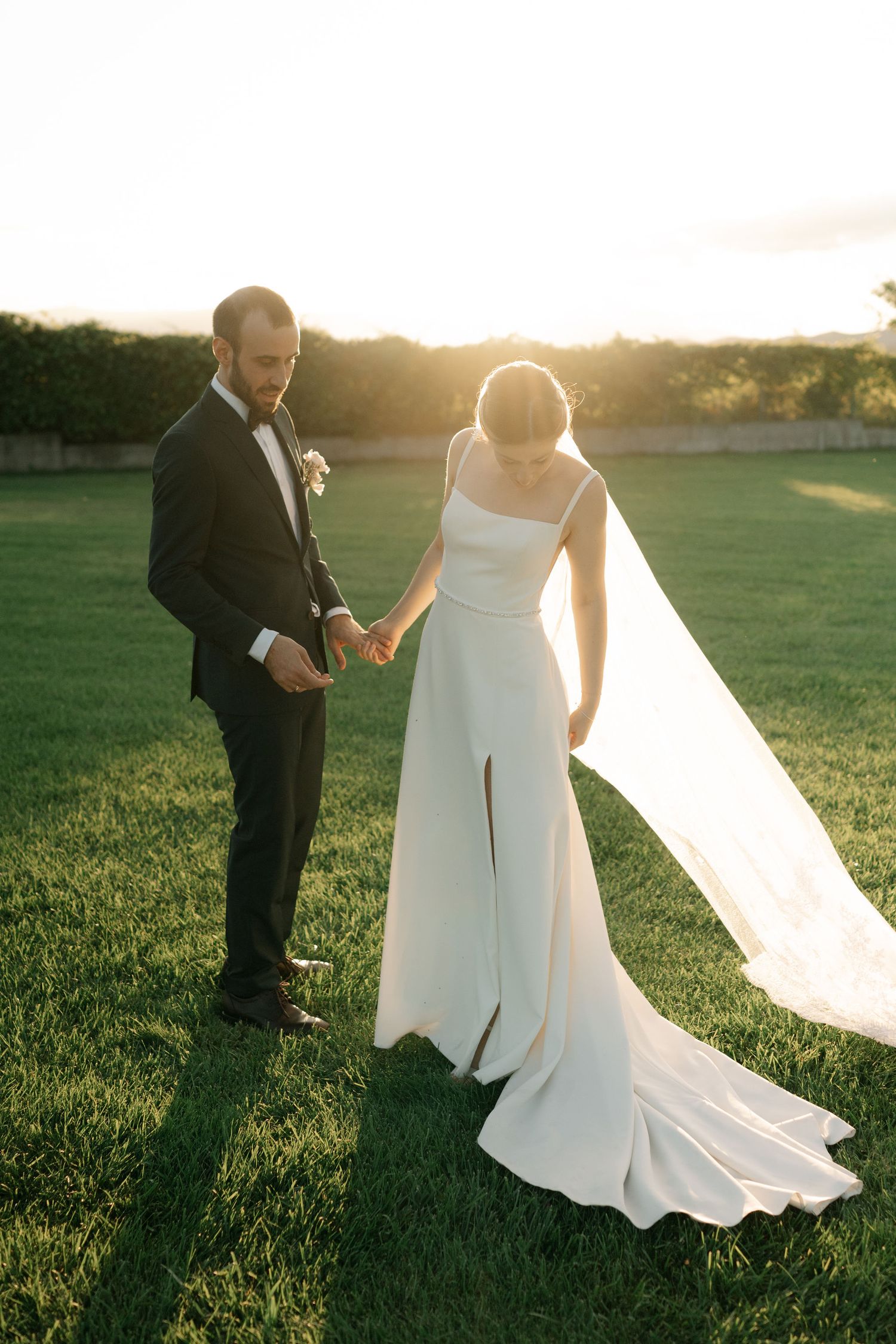 A couple in wedding attire stands on a sunlit grassy field during golden hour, with the bride's dress flowing in the breeze.