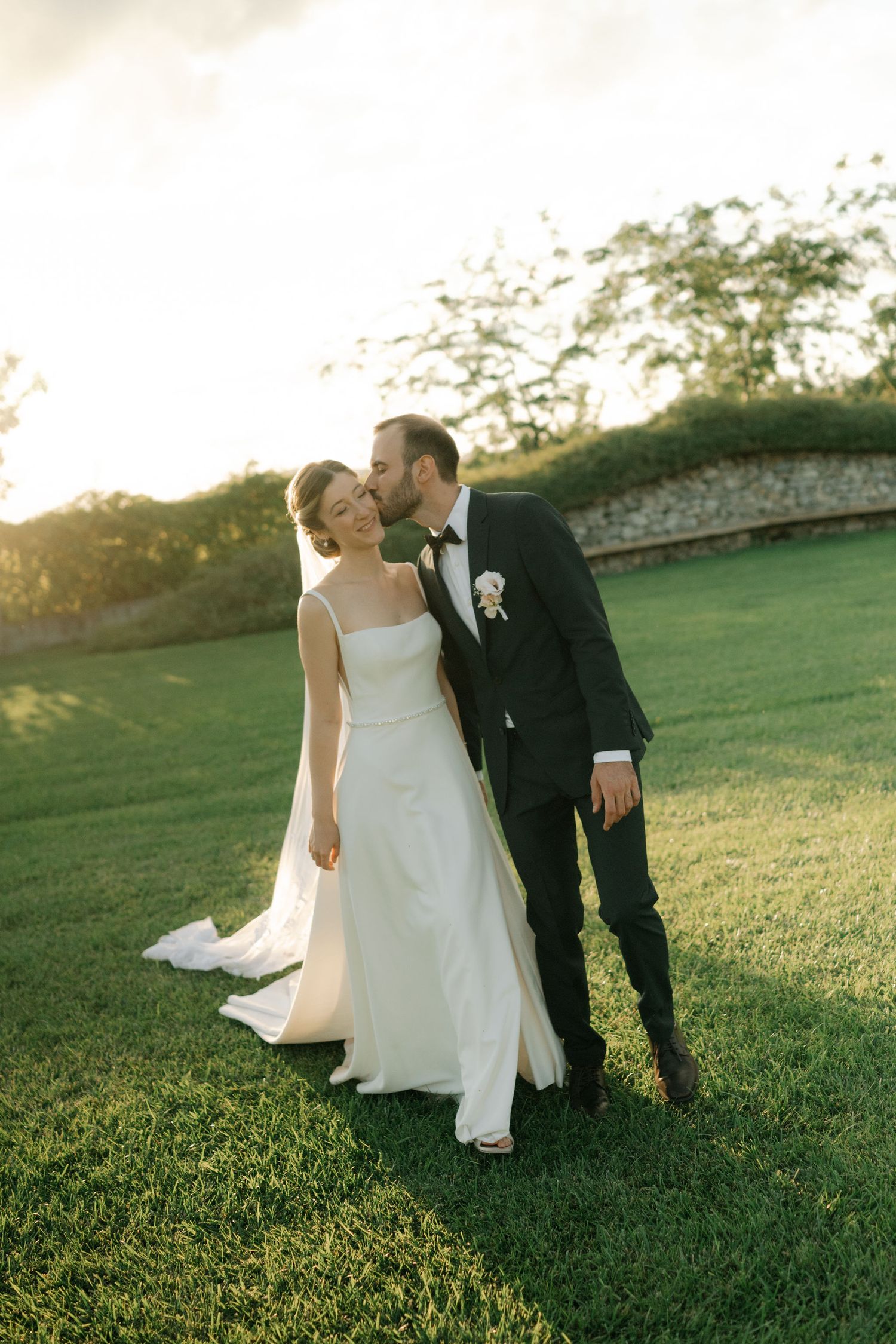 Newlywed couple shares intimate moments on a sunlit grassy hillside during golden hour wedding photography session.