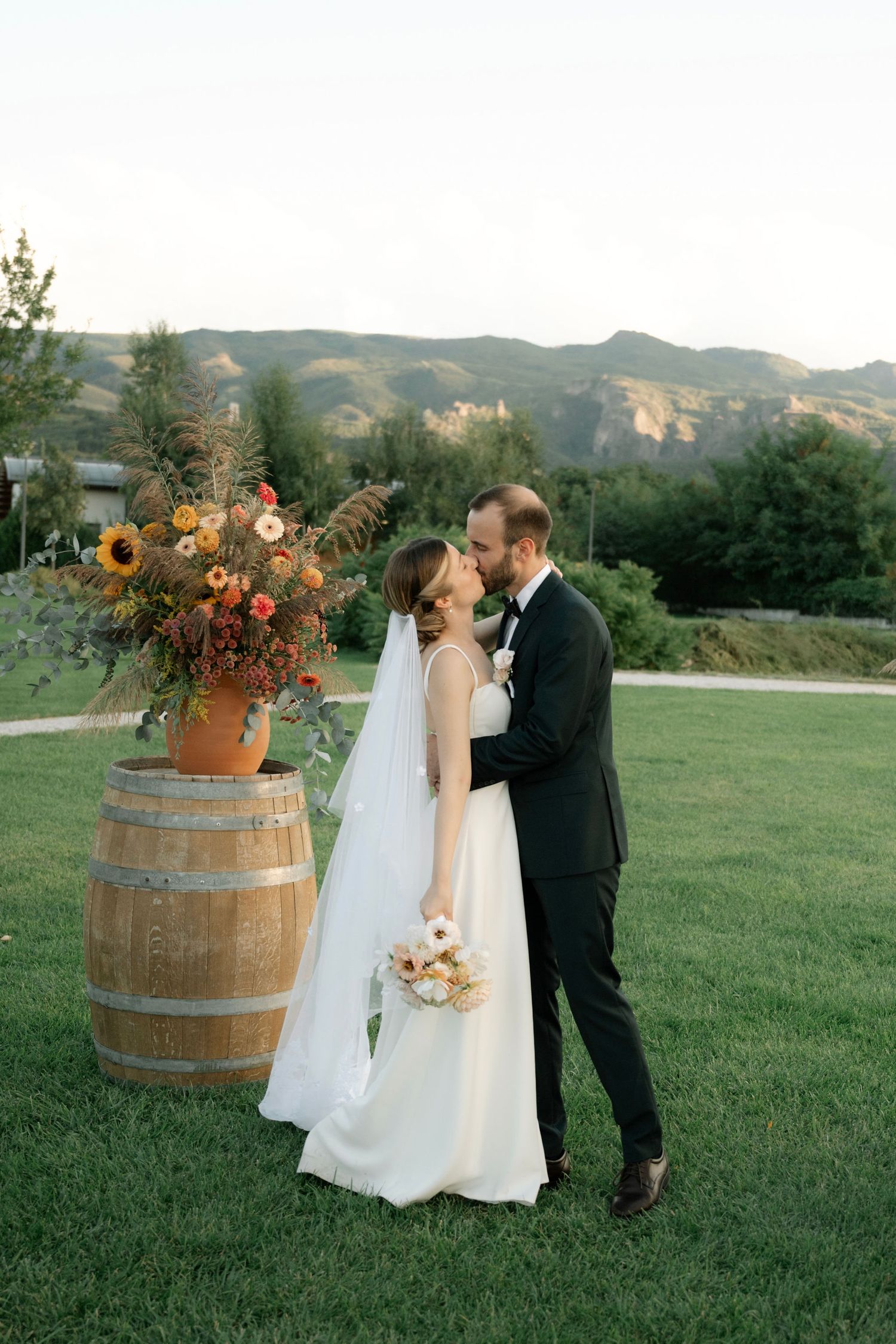 A romantic outdoor wedding scene with a wine barrel and sunflower arrangement against a mountain backdrop.