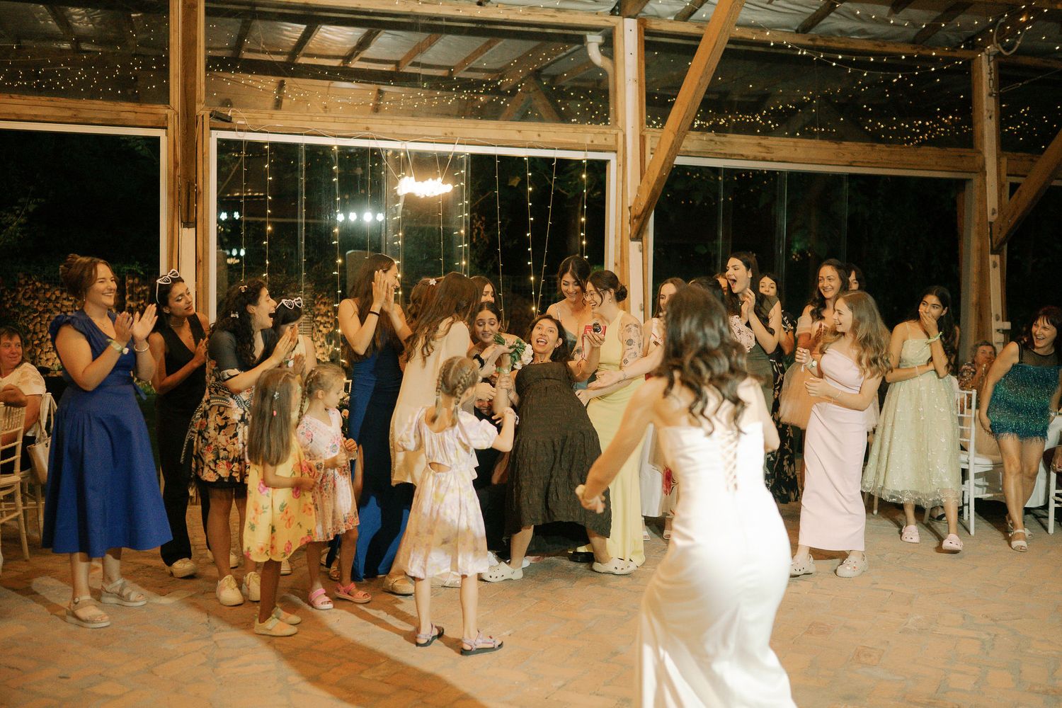 Wedding guests dance together on a rustic wooden dance floor decorated with string lights during an evening reception.
