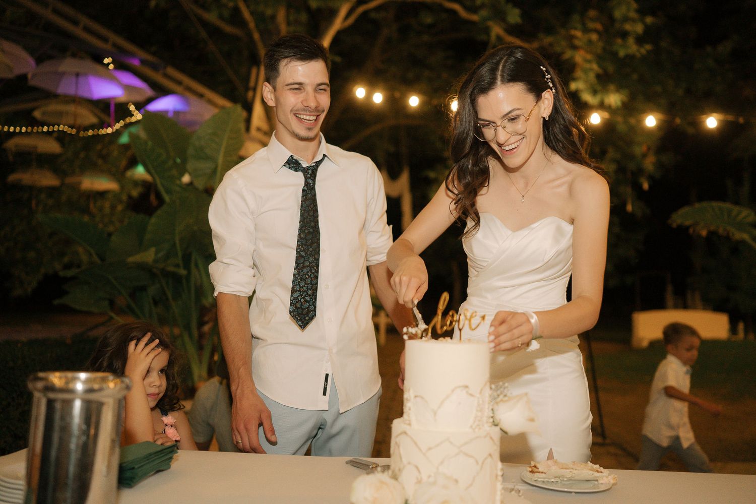 Newlyweds cut their wedding cake together at an outdoor evening reception with string lights glowing in the background.