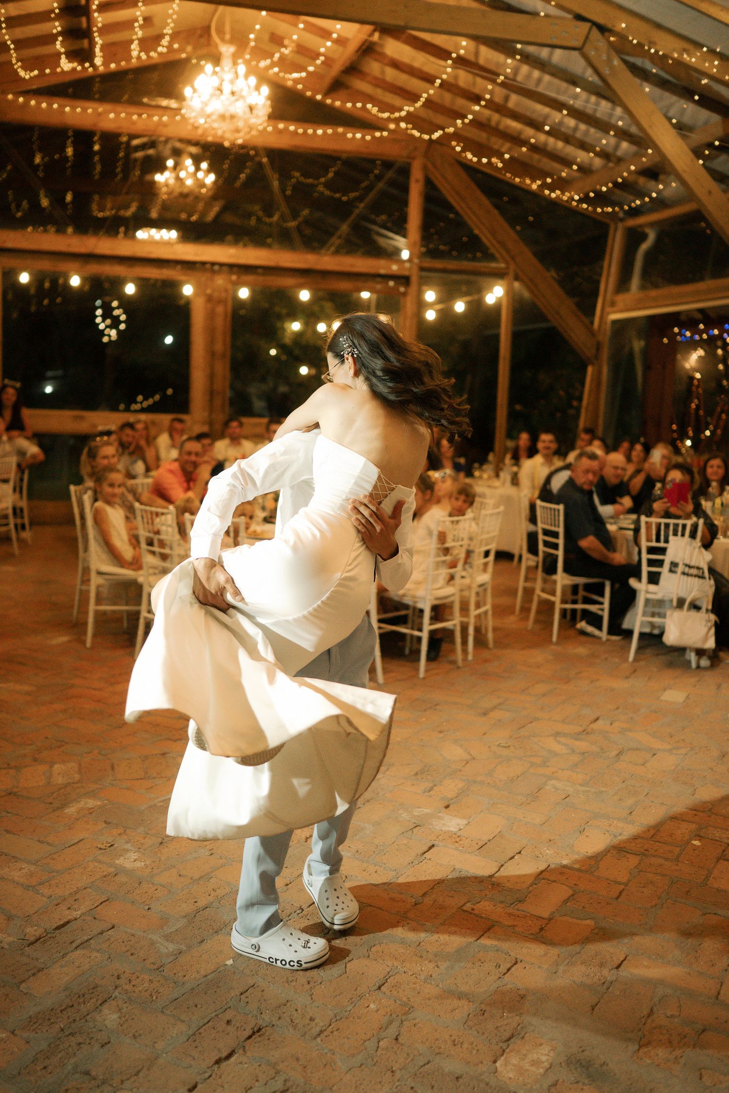 A couple dances together during a wedding reception in a rustic venue decorated with twinkling string lights and chandeliers.
