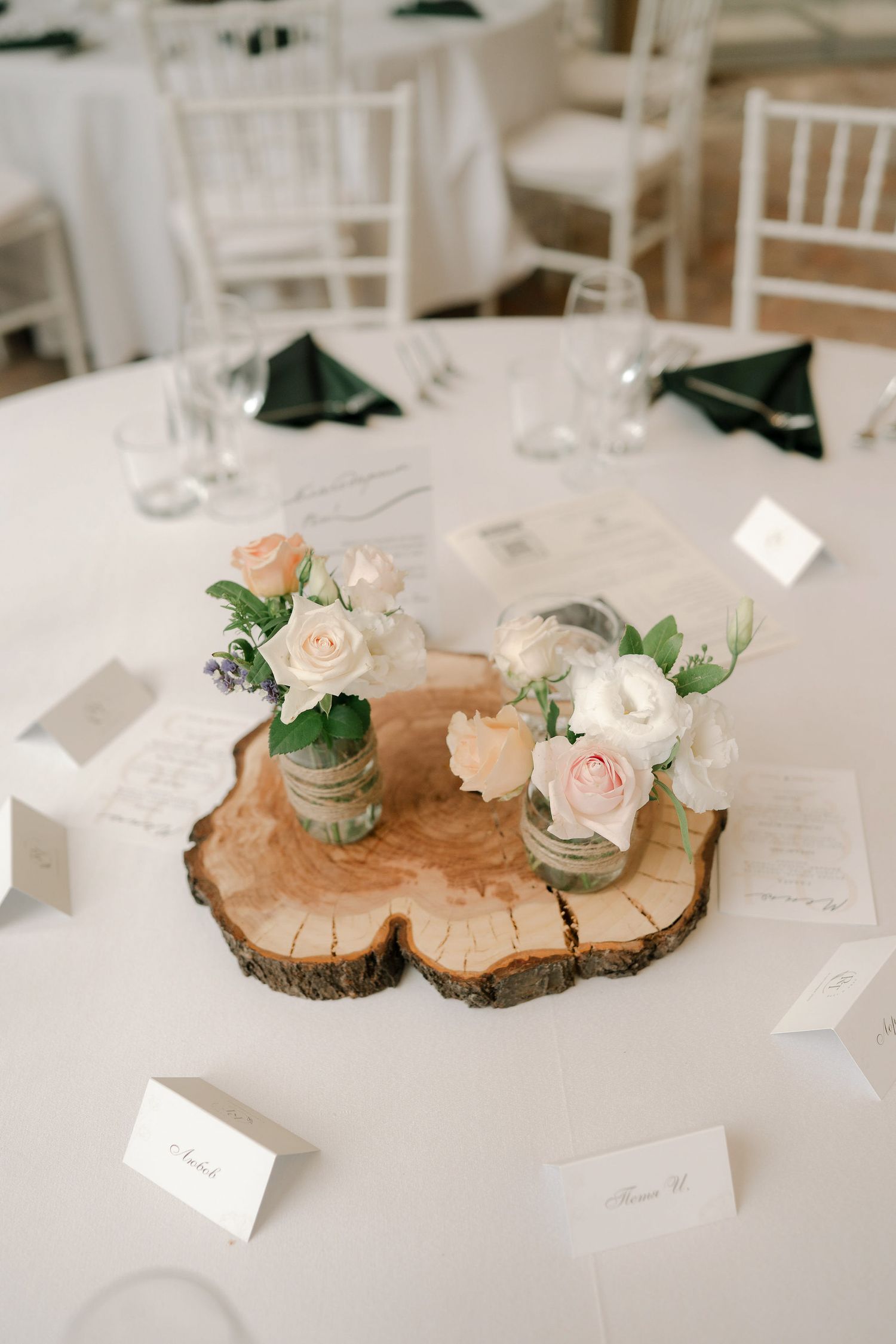 Rustic wedding table decor featuring wood slice centerpieces with pink roses and mason jars on white tablecloths.